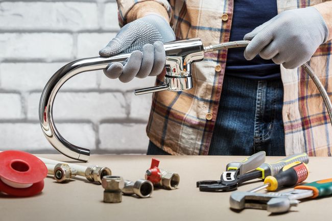 A person in gloves holding a kitchen faucet over a table with plumbing tools and hardware against a brick wall.