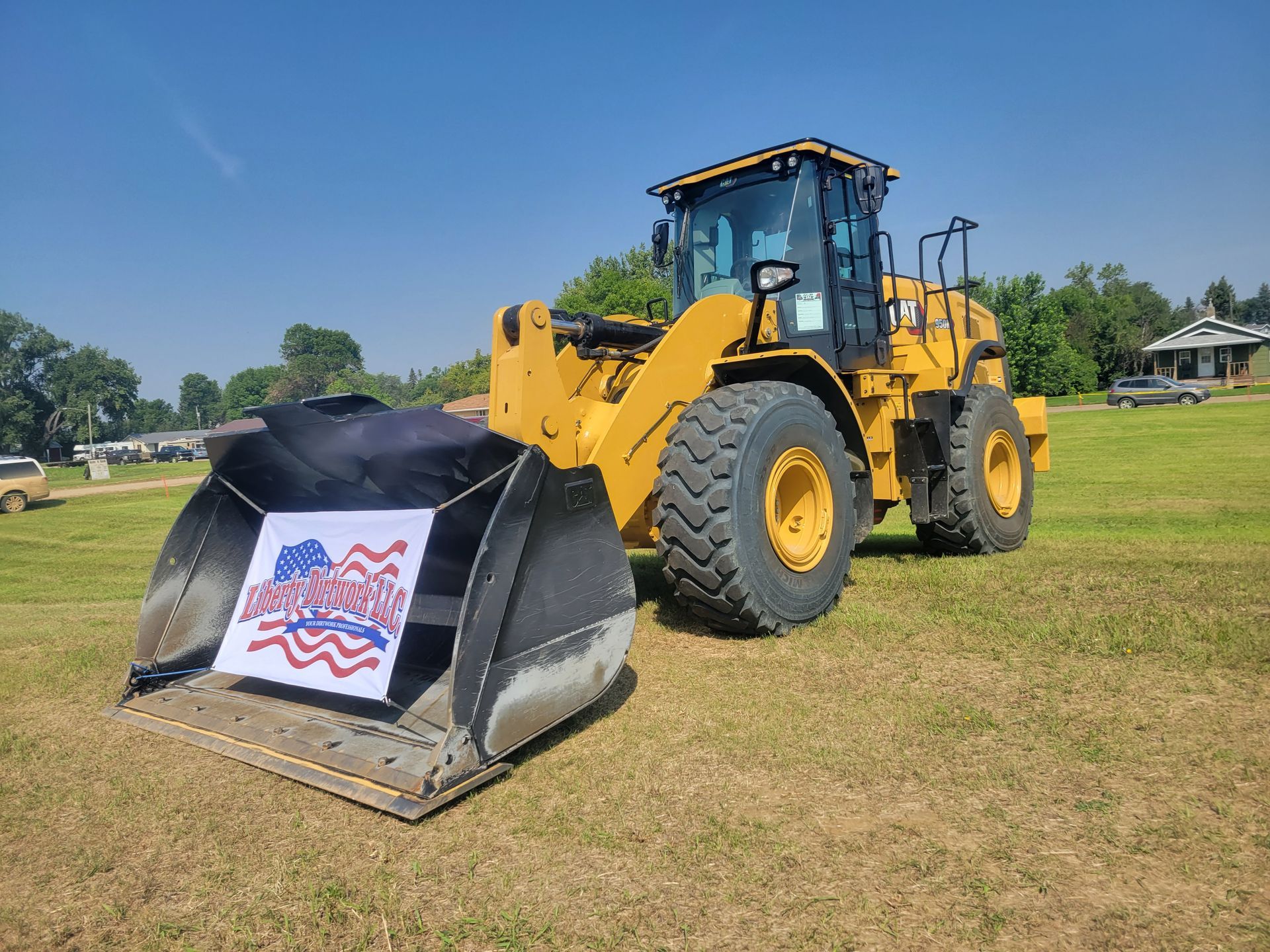 Yellow construction loader with a patriotic sign on its bucket, parked on a grassy field under a blue sky.