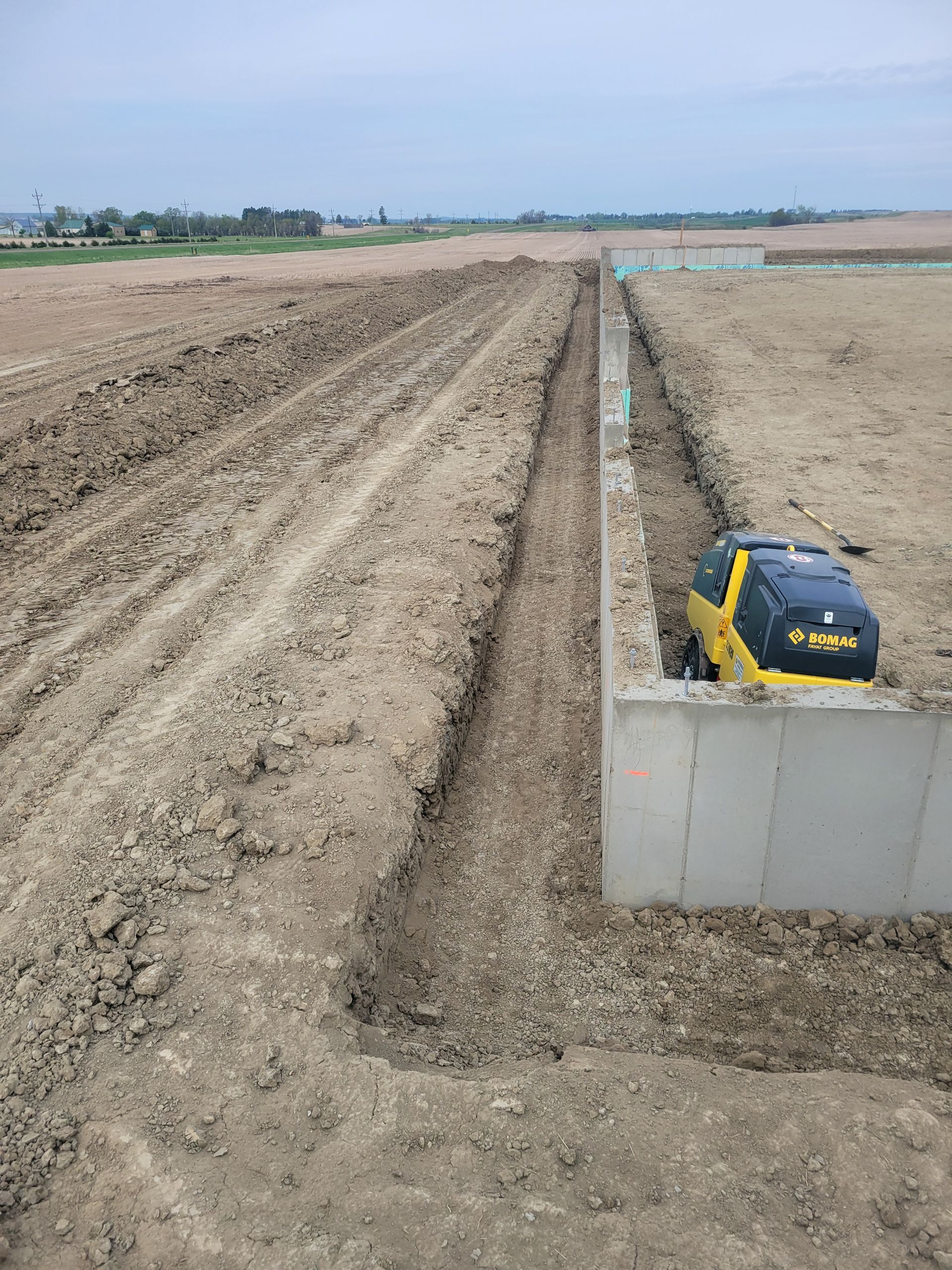 Construction site: Trenches dug in brown dirt with a yellow and black compactor on a concrete foundation.