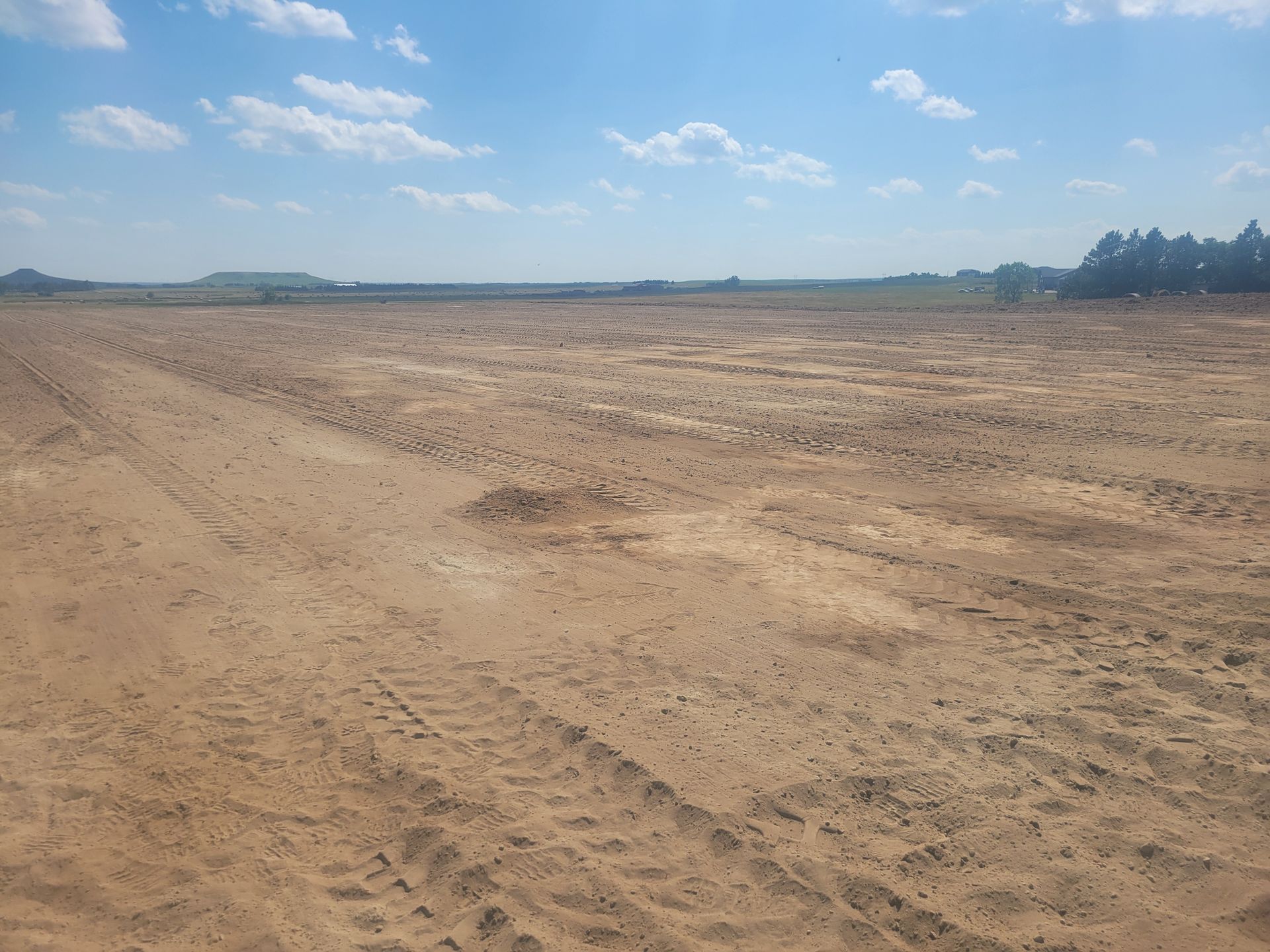 Brown field under a blue sky with scattered clouds. Vehicle tracks are visible.