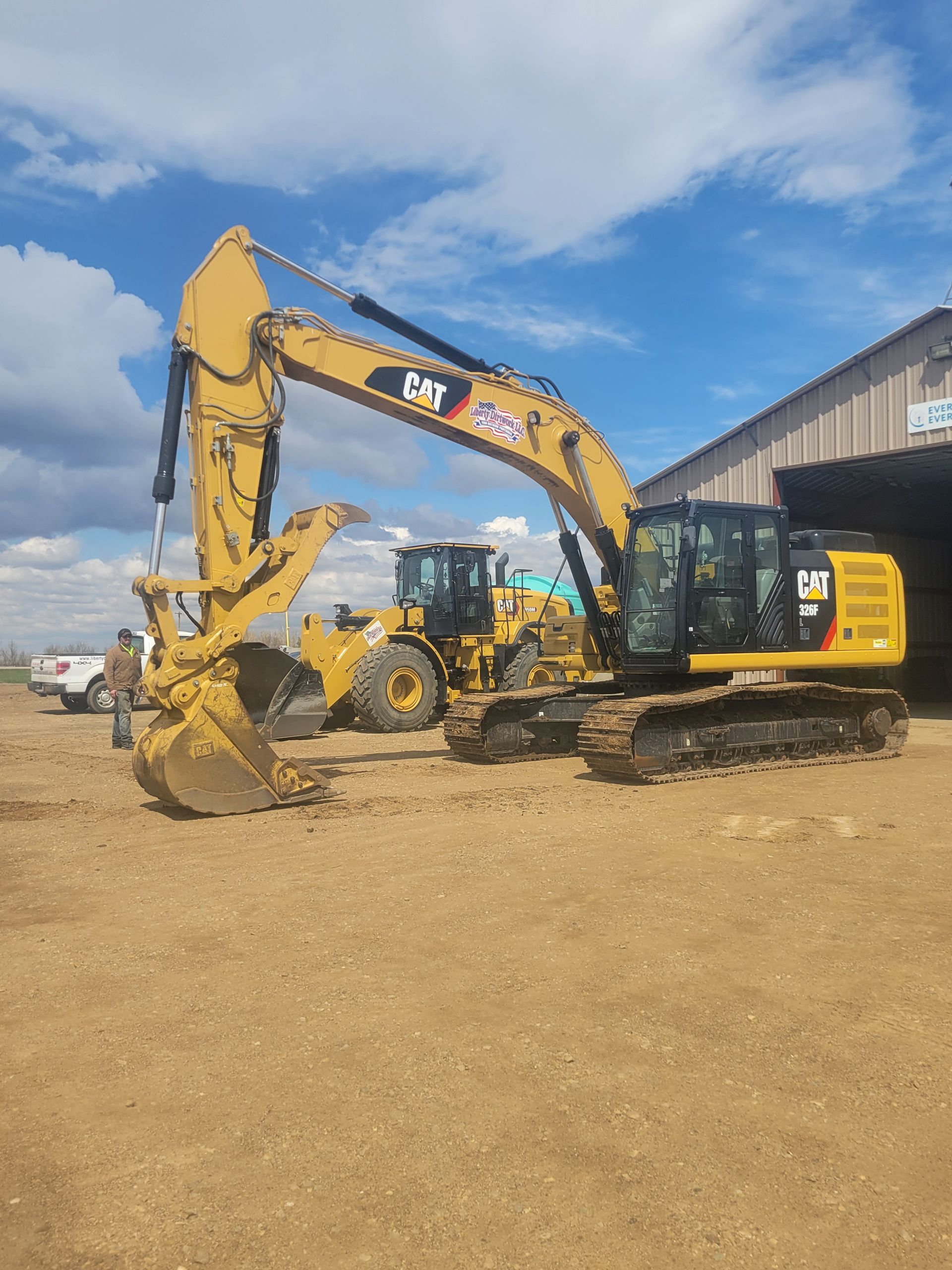 Yellow Caterpillar excavator with long arm, parked on dirt with other equipment under a blue sky.