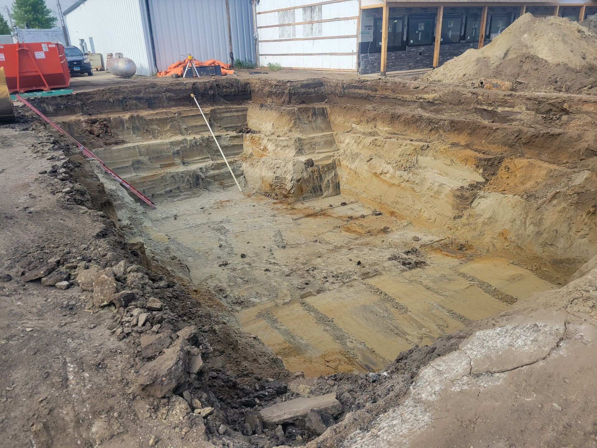 Excavated rectangular foundation pit, stepped sides, beige soil. Building and equipment visible in the background.