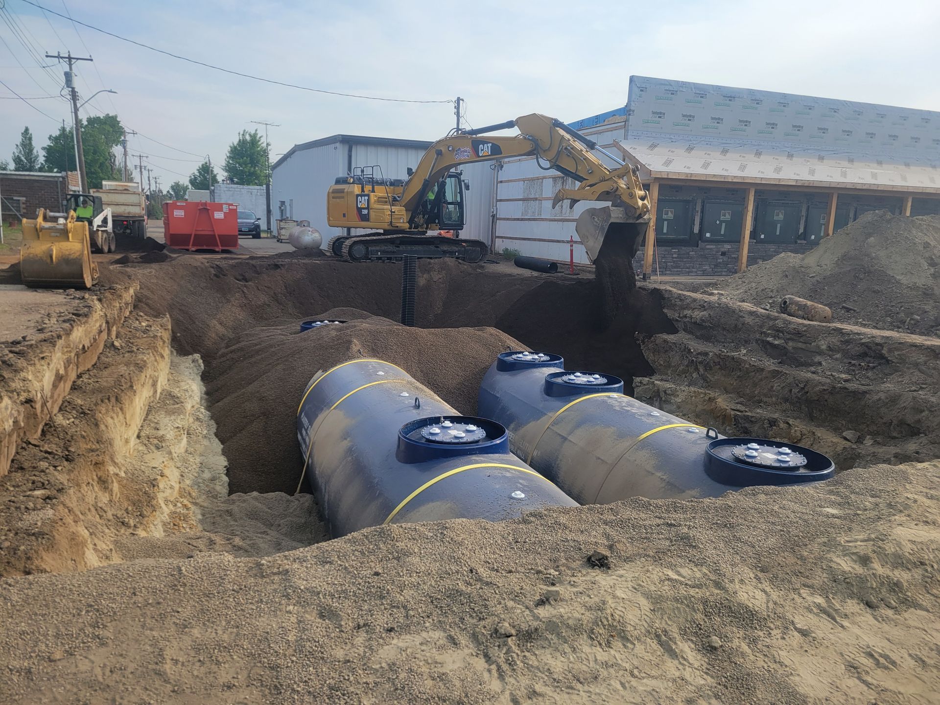 Excavator filling a trench with gravel to bury two large, blue tanks. Construction site.