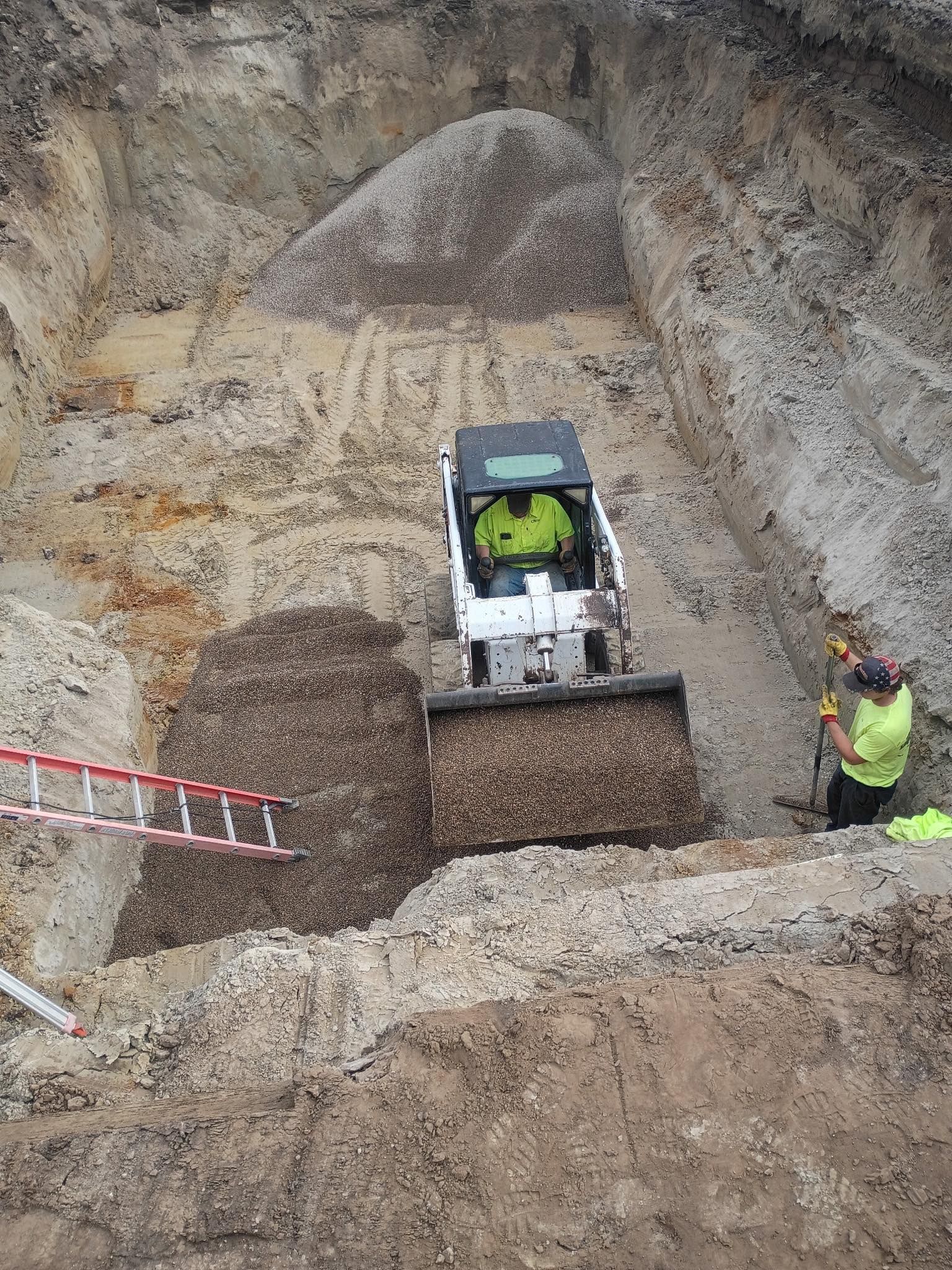 Bobcat compact track loader operating in a construction excavation. Worker with shovel on right, ladder on left.