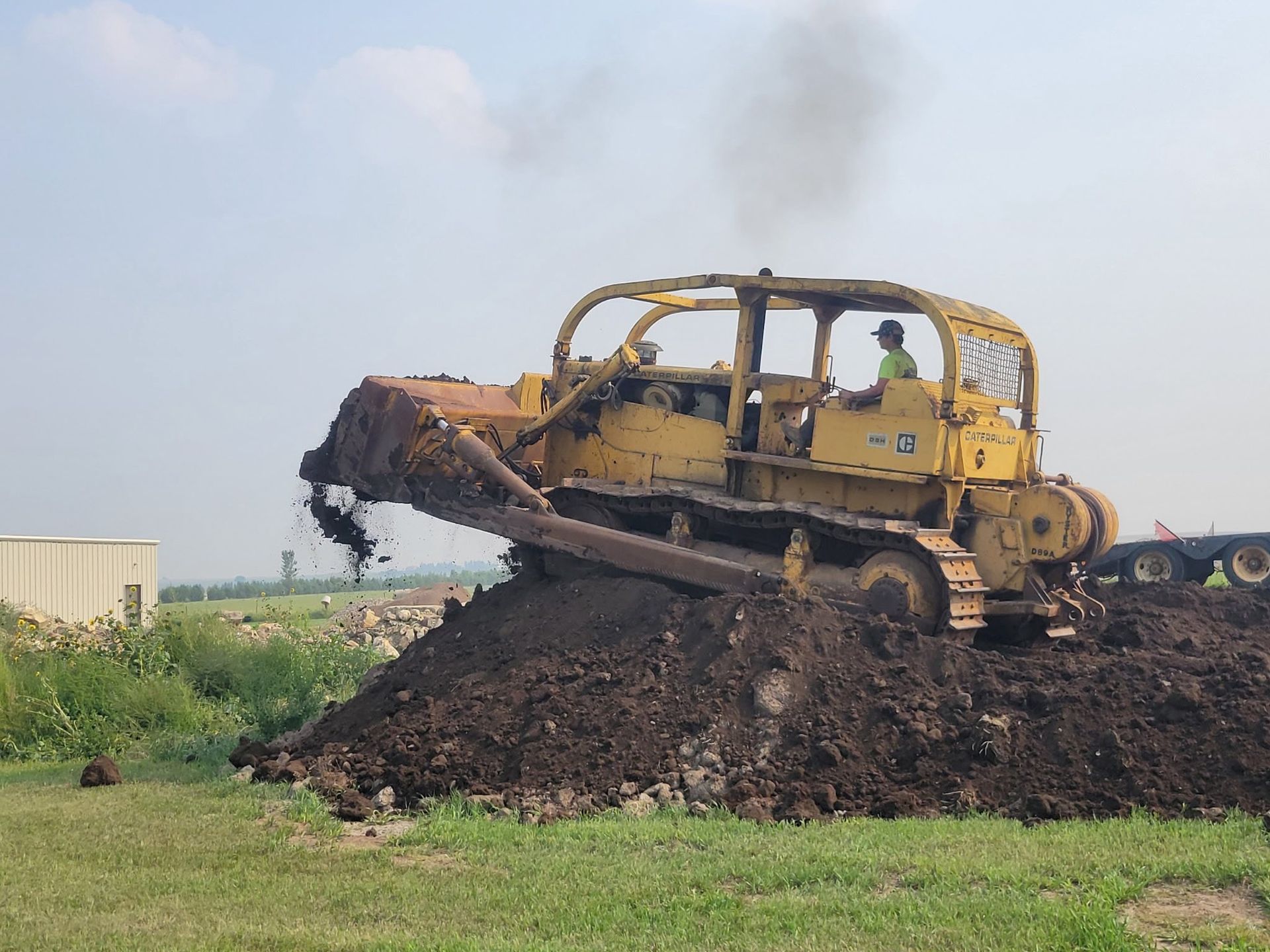 Yellow bulldozer pushing dark soil on a grassy field under a hazy sky.