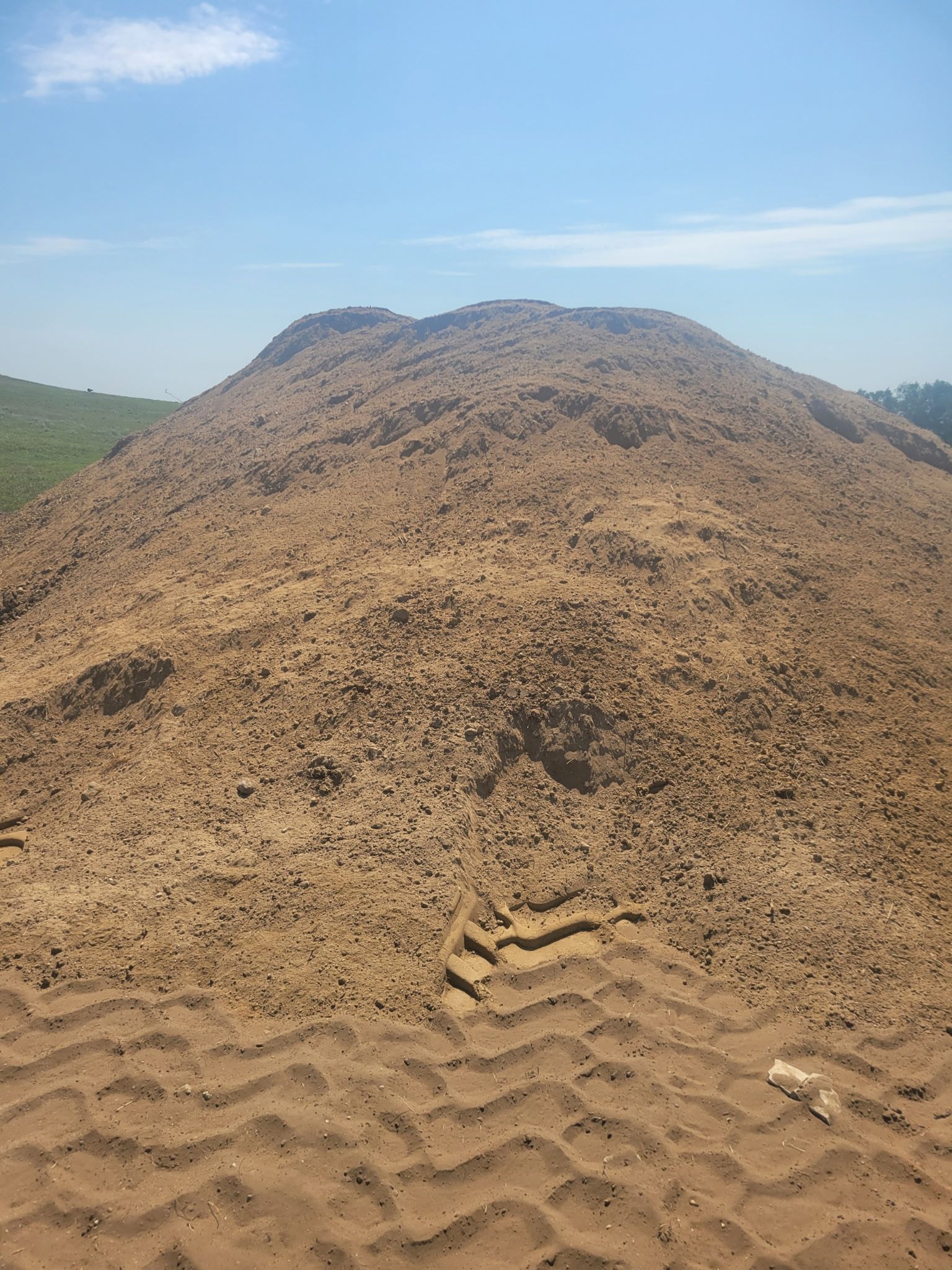 Large pile of tan sand with tire tracks in the foreground, against a blue sky.