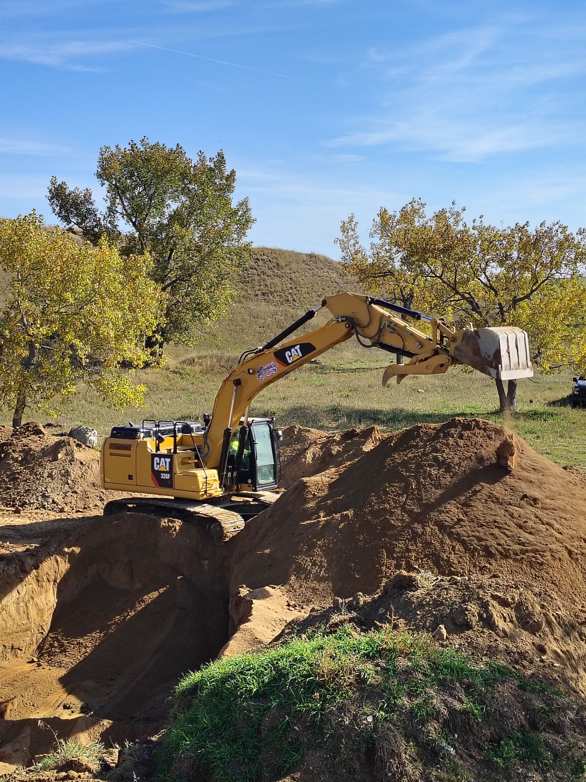 Yellow excavator digging in brown soil under a blue sky, trees in the background.