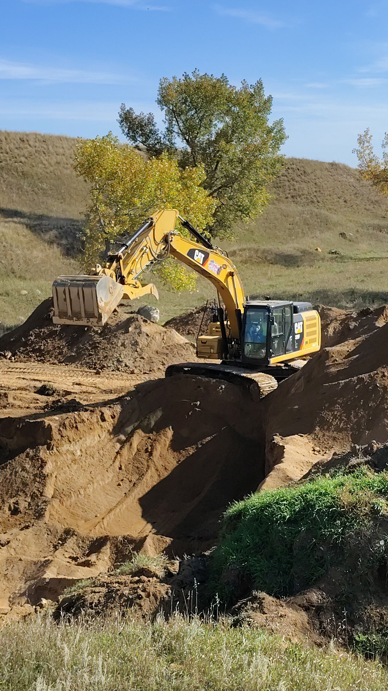Yellow excavator digging into a brown embankment with a tree on top, under a blue sky.