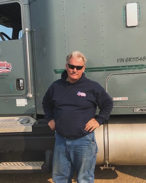 Man in sunglasses stands next to a green semi-truck; hands on hips.
