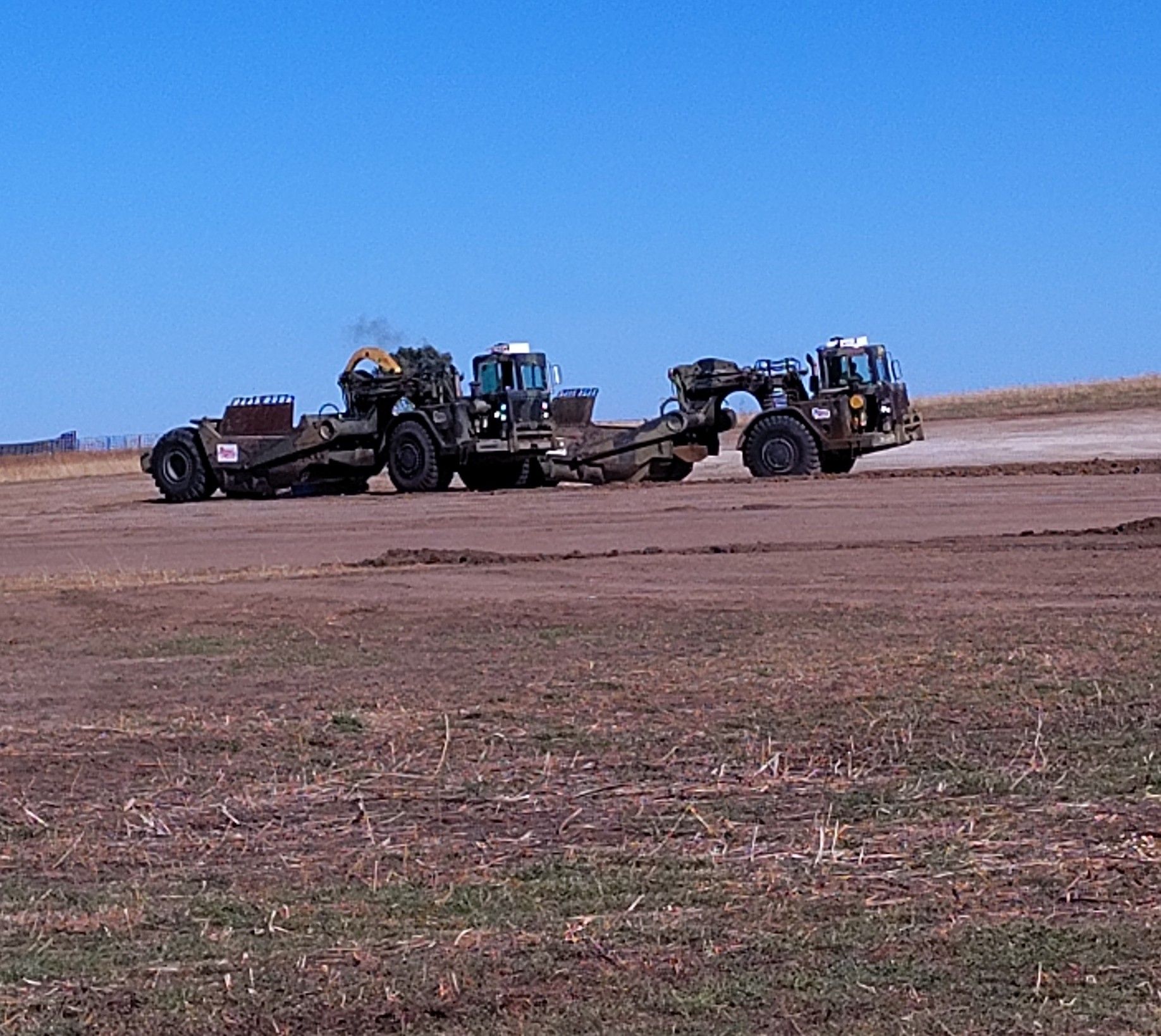 Two large earthmovers working on a field, moving dirt. Blue sky.