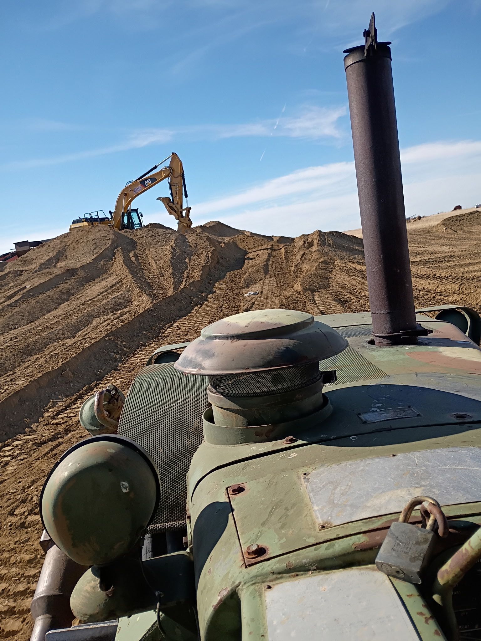 An old green tractor in the foreground with an excavator working on a dirt pile in the background.
