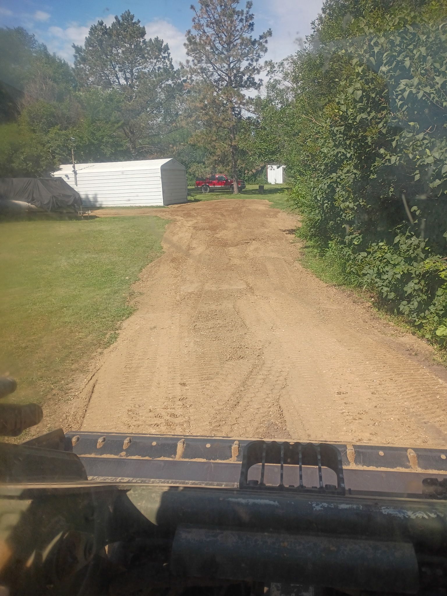 A dirt driveway leading towards a red truck with a white shed on the left and trees on the right.