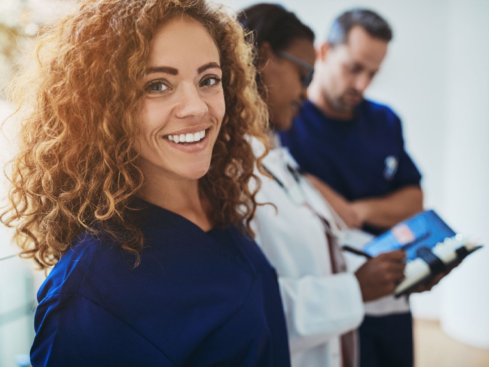 A woman in a blue scrub is smiling in front of a group of doctors.