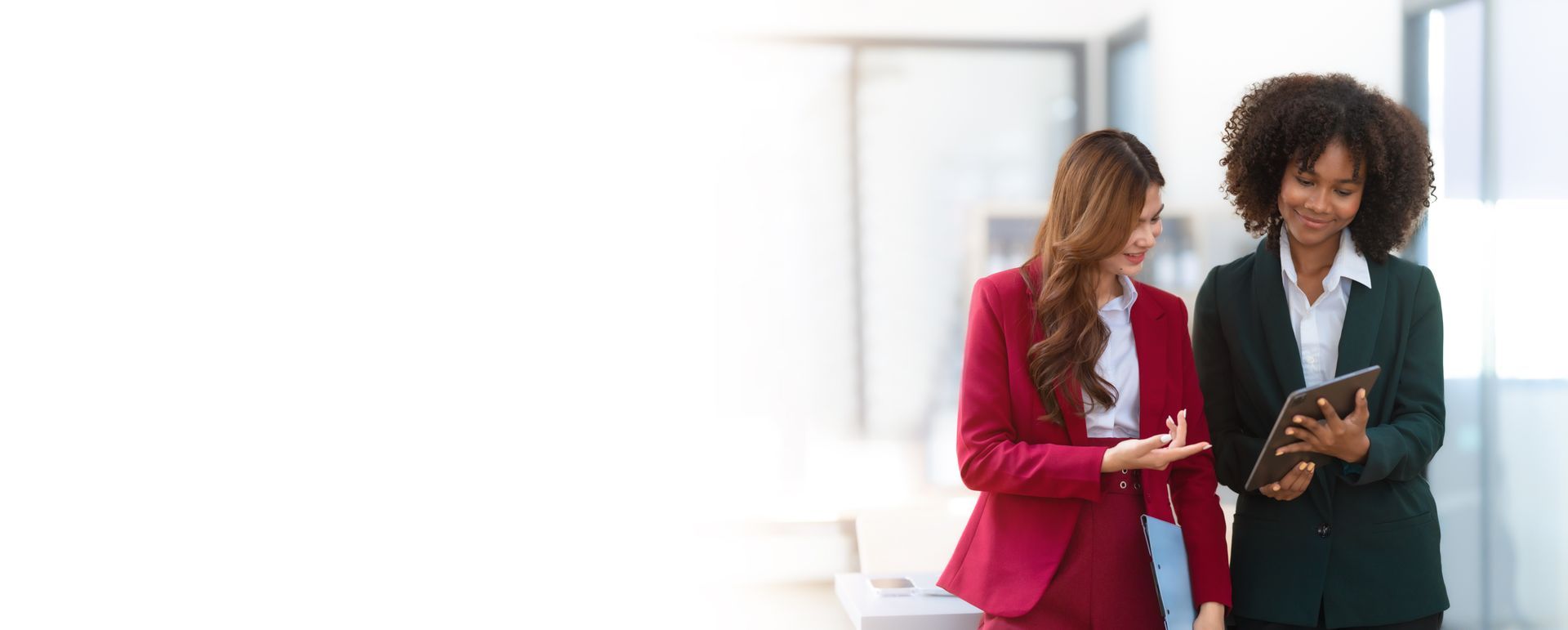 Two business women are standing next to each other in an office looking at a tablet.