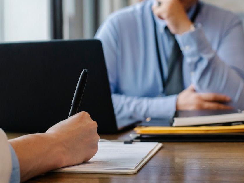 A man is sitting at a desk with a laptop and a woman is writing in a notebook.