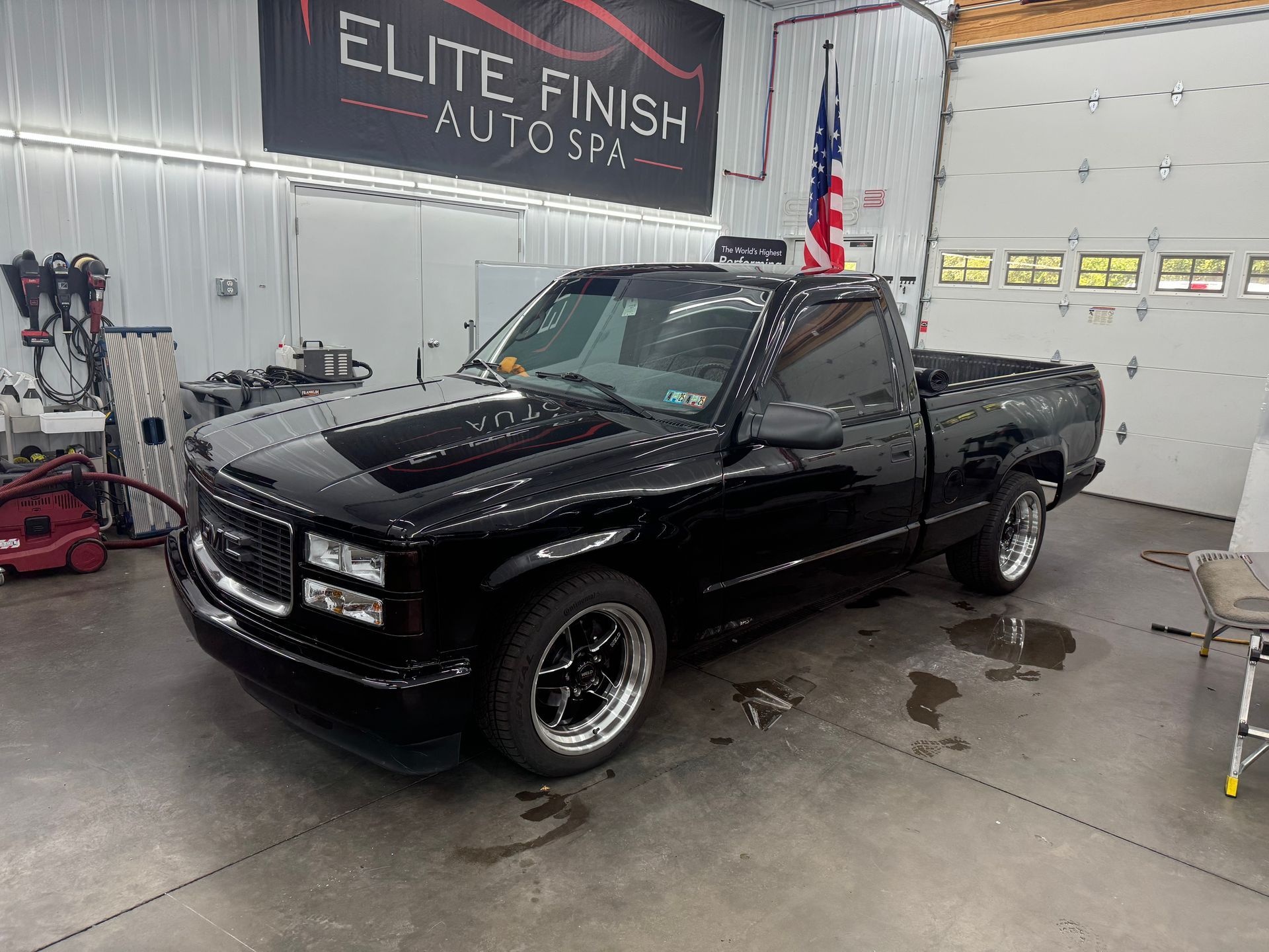 A black truck is parked in a garage next to an american flag.