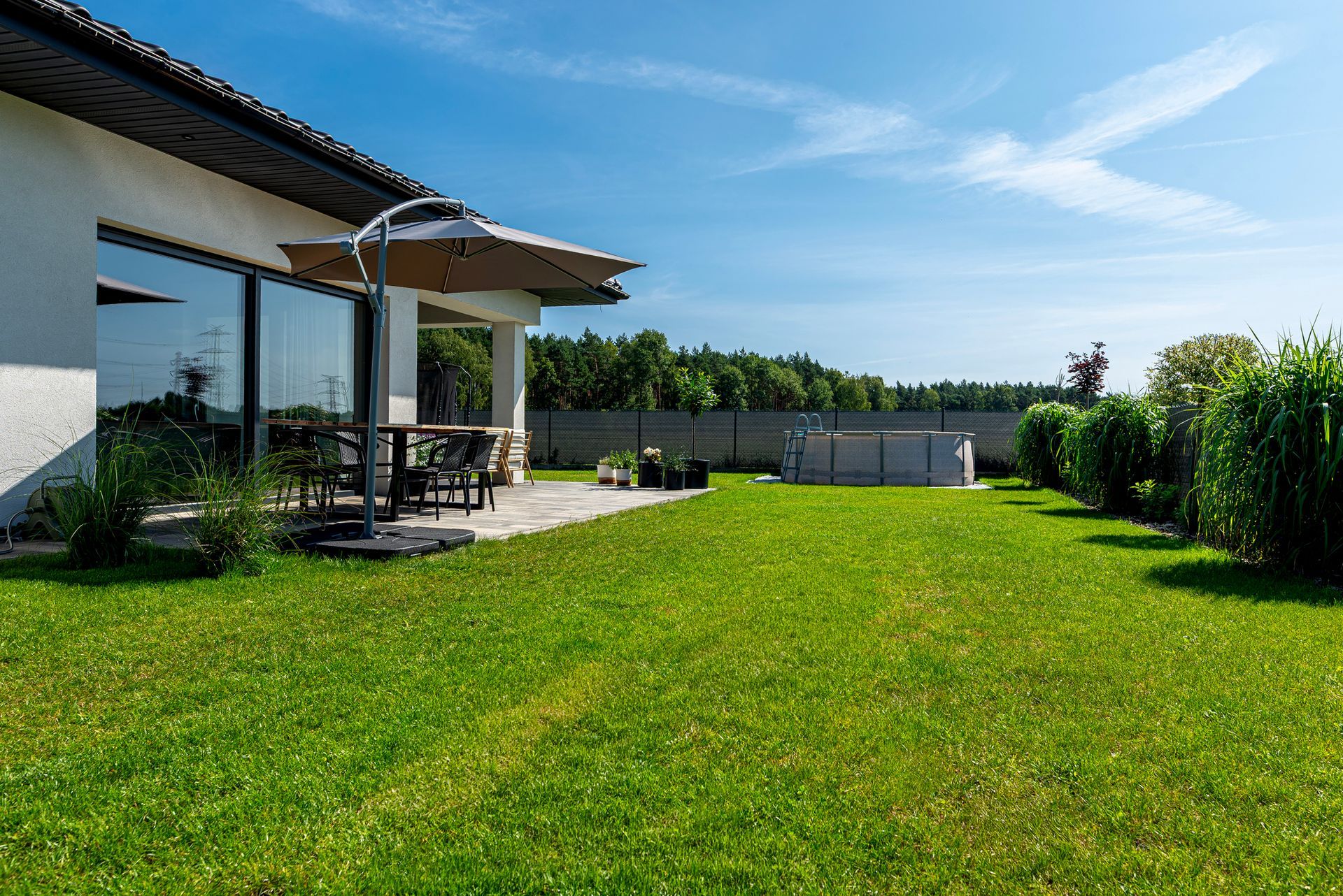 Prato e patio di una casa in una giornata di sole. Tavolo con sedie, piscina e alberi sullo sfondo.