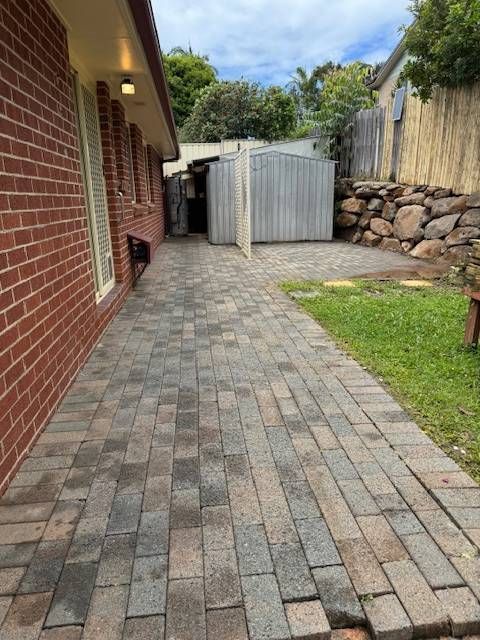 Brick pathway alongside a red brick house, leading to a metal shed in a backyard with a retaining wall.