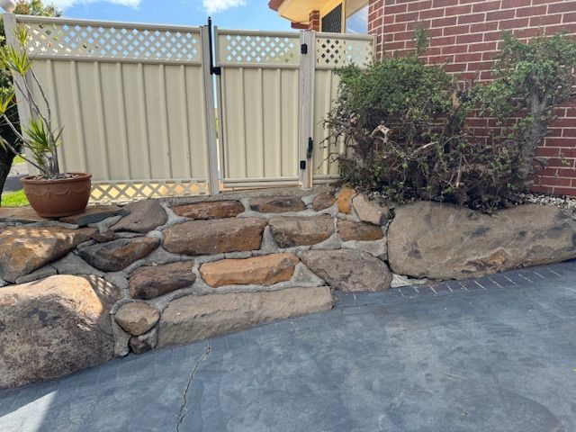 Stone steps lead to a gate beside a red brick building and beige fence. A potted plant sits on the left.