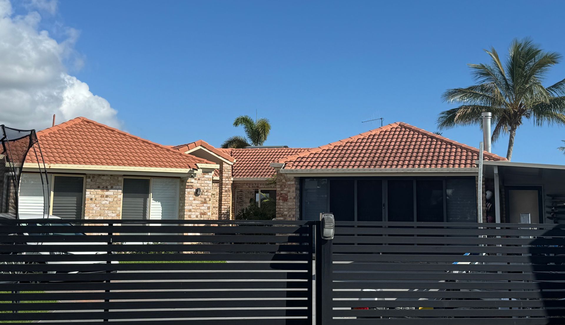 House with red tile roof behind a black slat fence on a sunny day with palm tree visible.