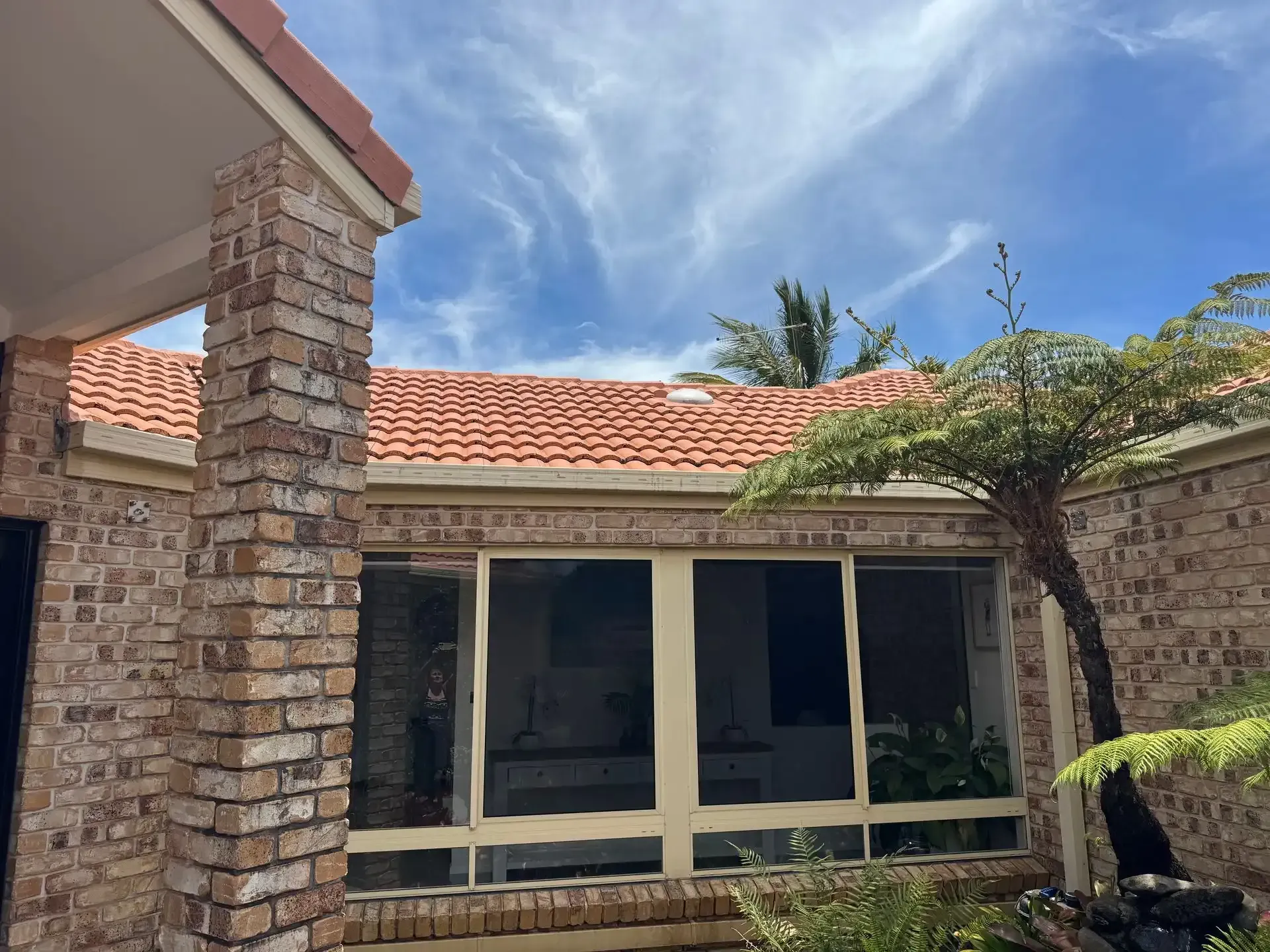 Brick home with tiled roof, large windows, and a tree, under a partly cloudy blue sky.