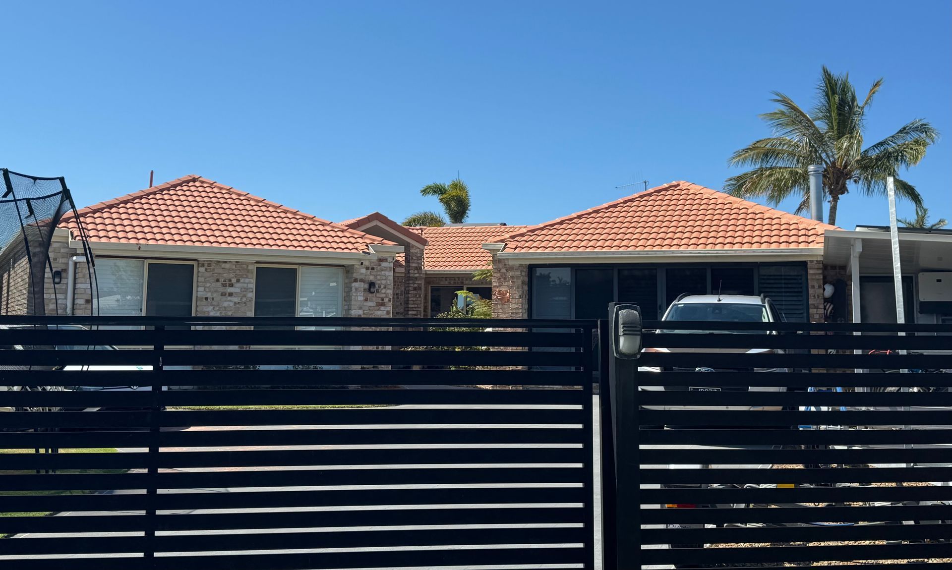 A brick home with orange tile roofs, behind a black fence. A car is visible. Palm tree nearby.