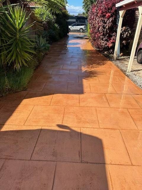 Brick-patterned orange driveway leading to a car, flanked by greenery and a covered carport on a sunny day.