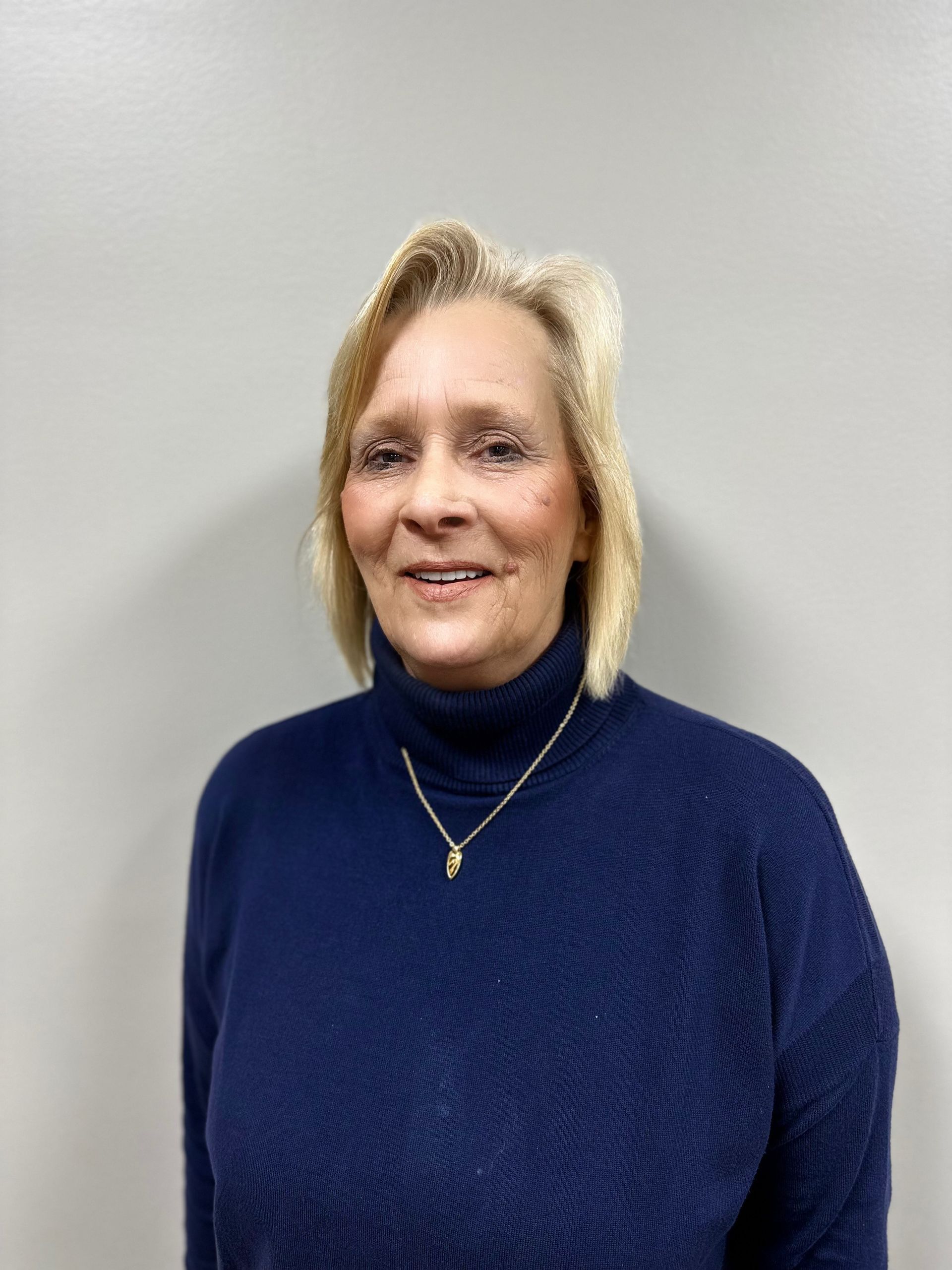 Woman in blue and white plaid shirt, hands clasped, standing in front of a gray backdrop.