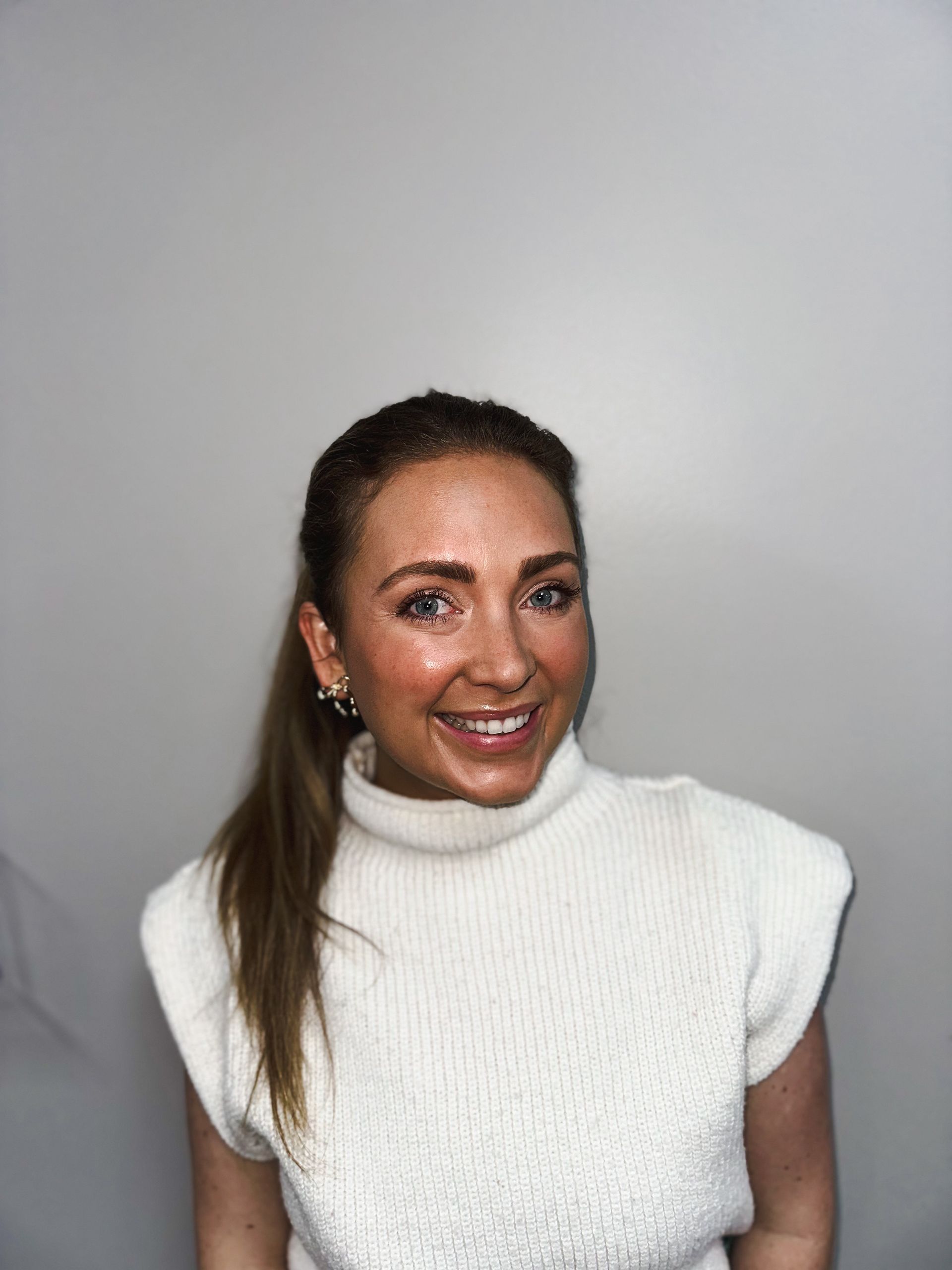 Woman with wavy blonde hair smiles, wearing a tan jumpsuit, standing in front of a gray backdrop.
