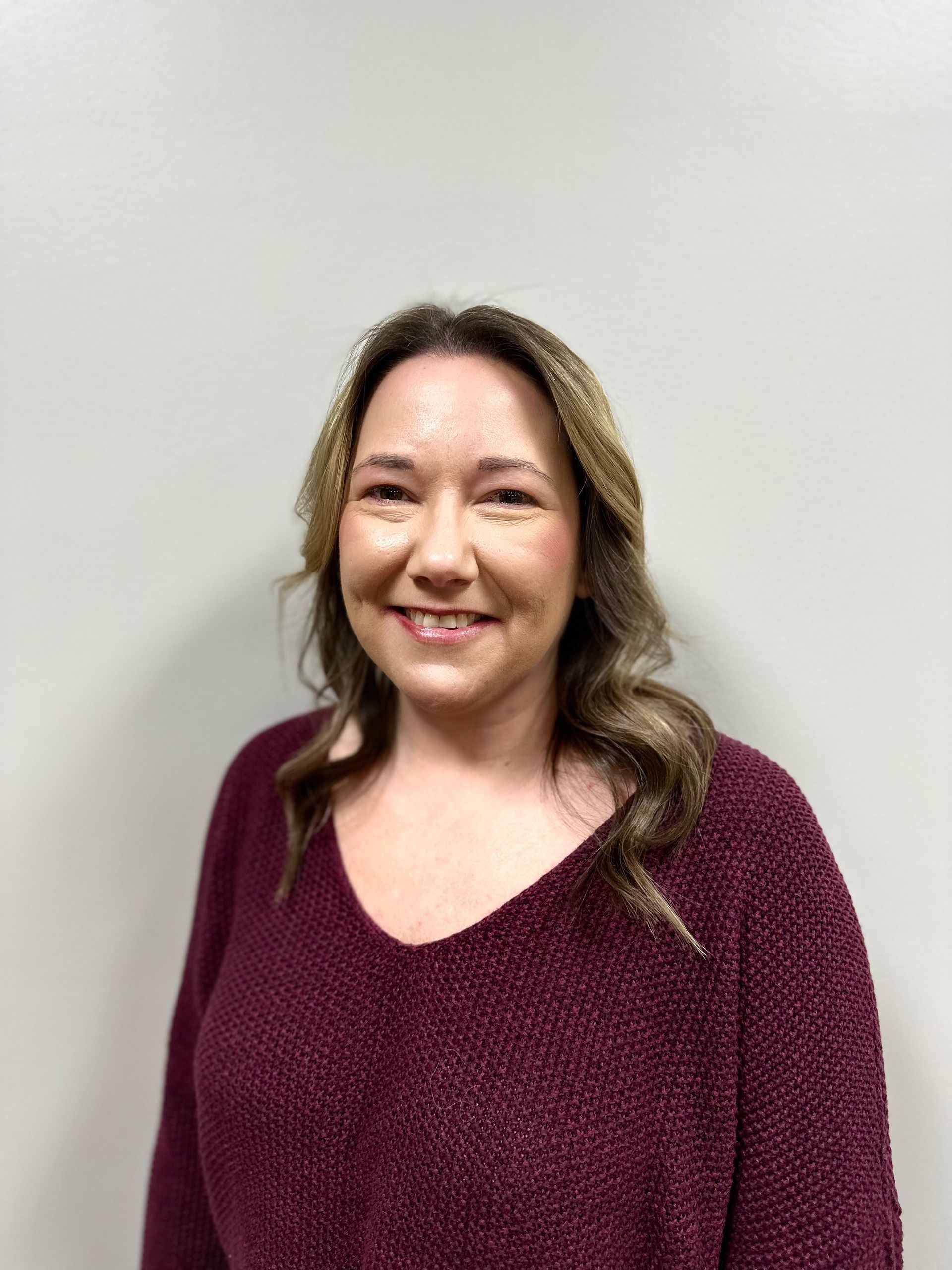 Woman with long, wavy blonde and brown hair wearing a blue sweater, smiling, arms crossed in front of a gray backdrop.