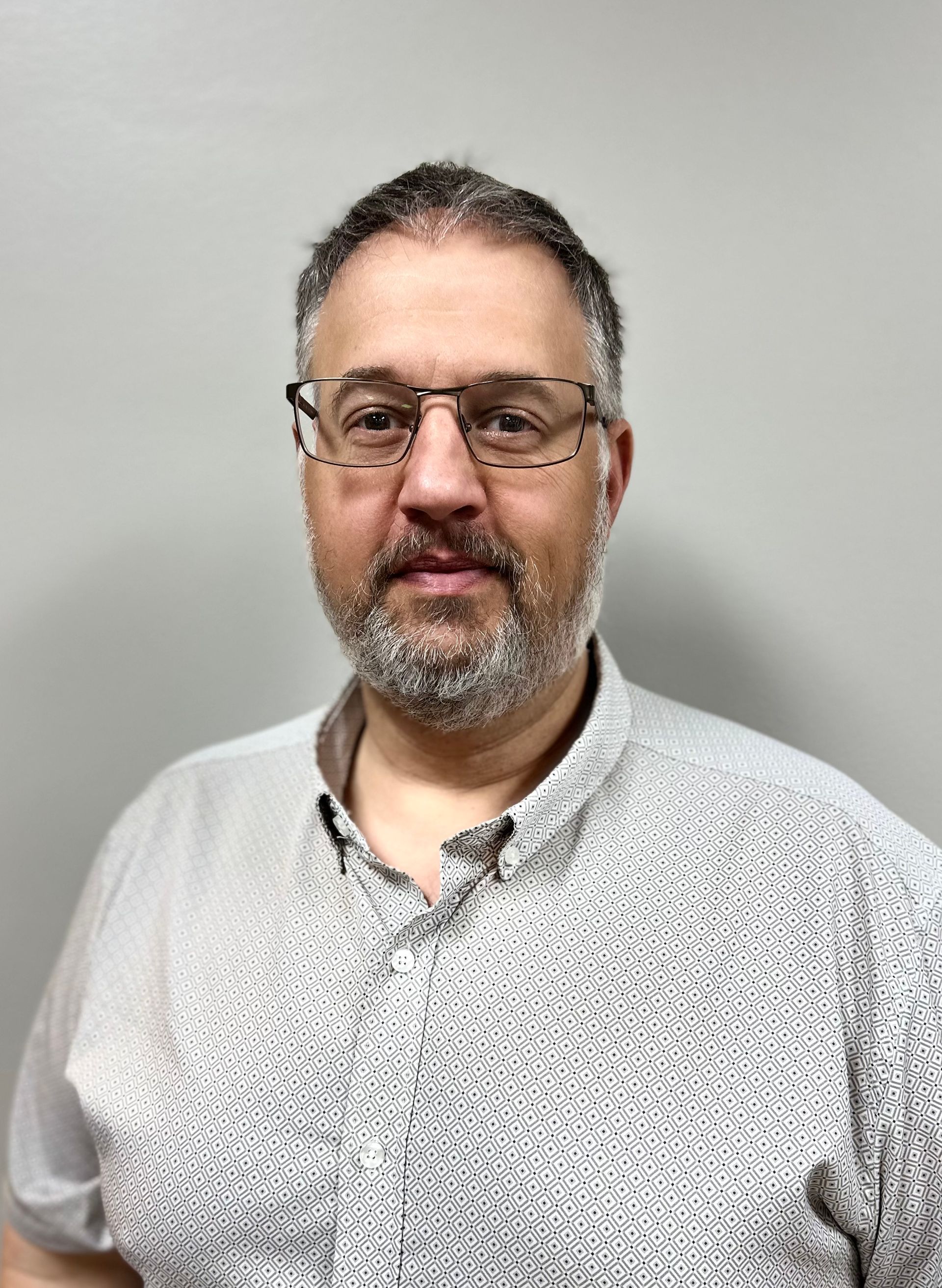 Man with glasses and short-sleeved blue shirt, arms crossed, stands against a gray textured backdrop.