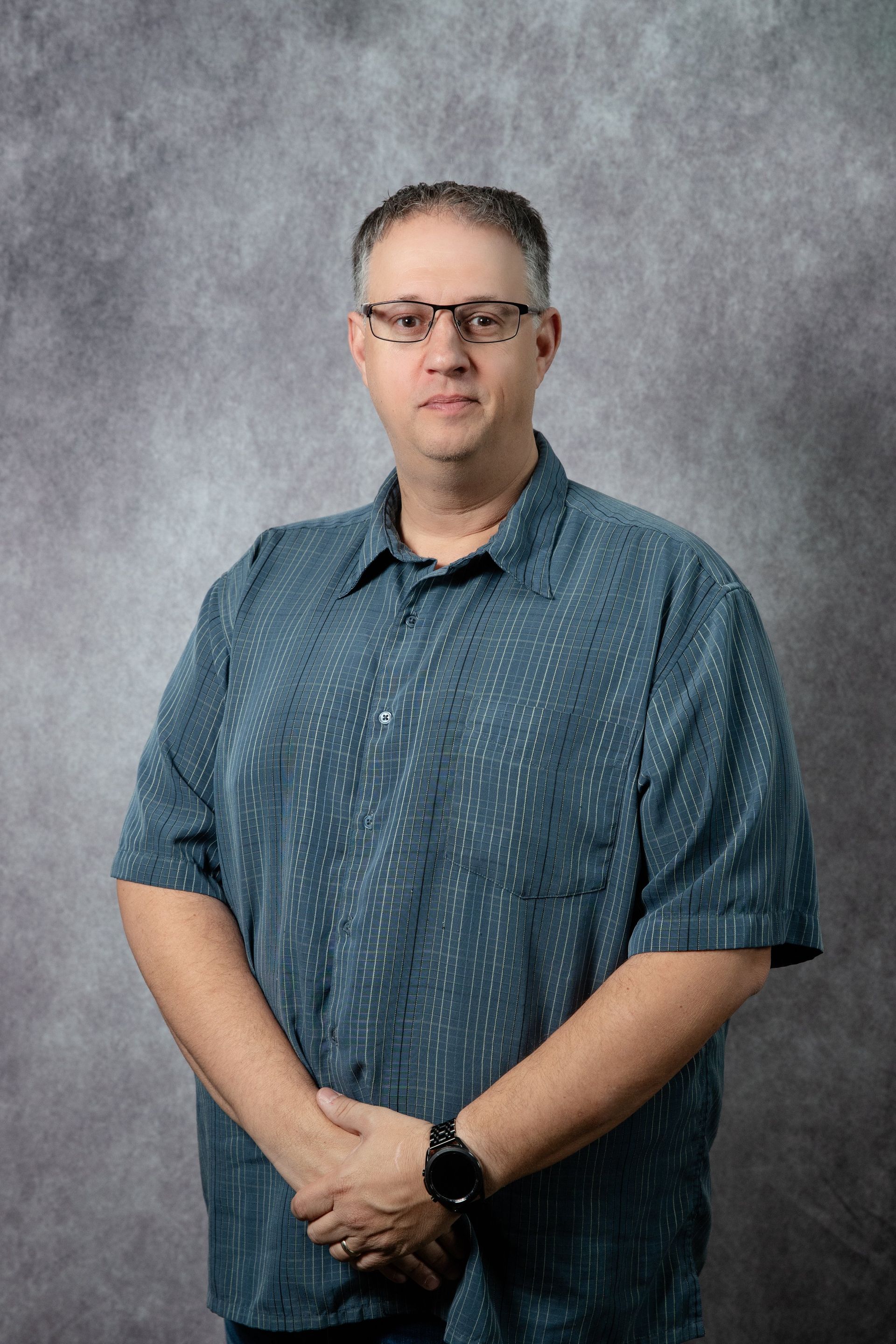 Man with glasses and short-sleeved blue shirt, arms crossed, stands against a gray textured backdrop.