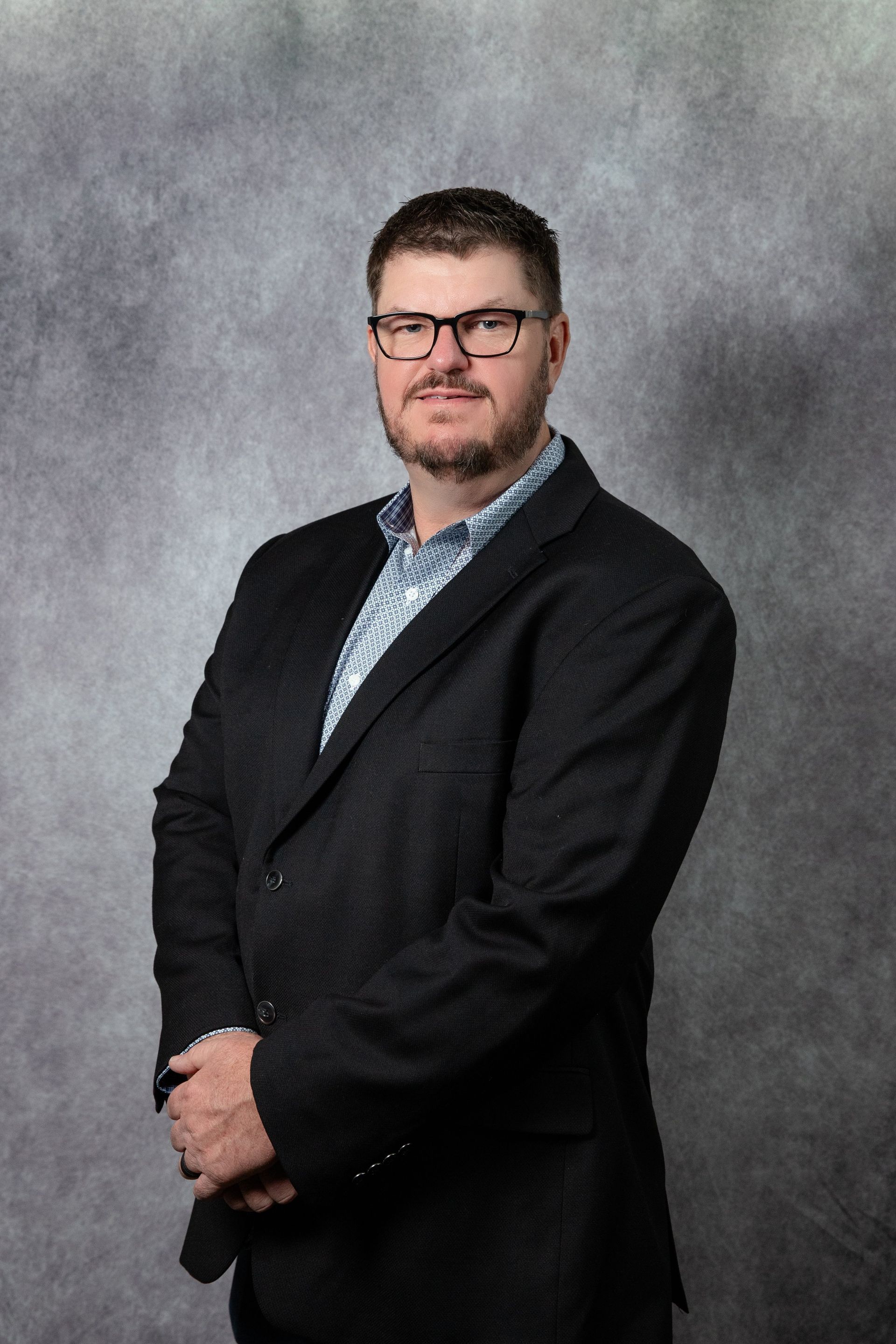 Man in suit and glasses, arms crossed, smiling. Gray background.