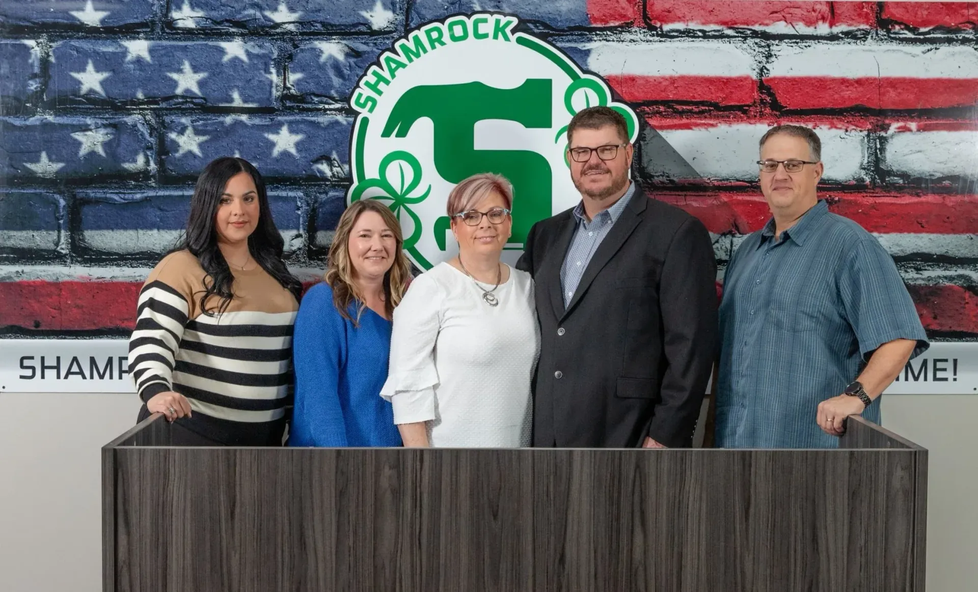 Group of five people standing behind a wooden counter with American flag backdrop and Shamrock logo.