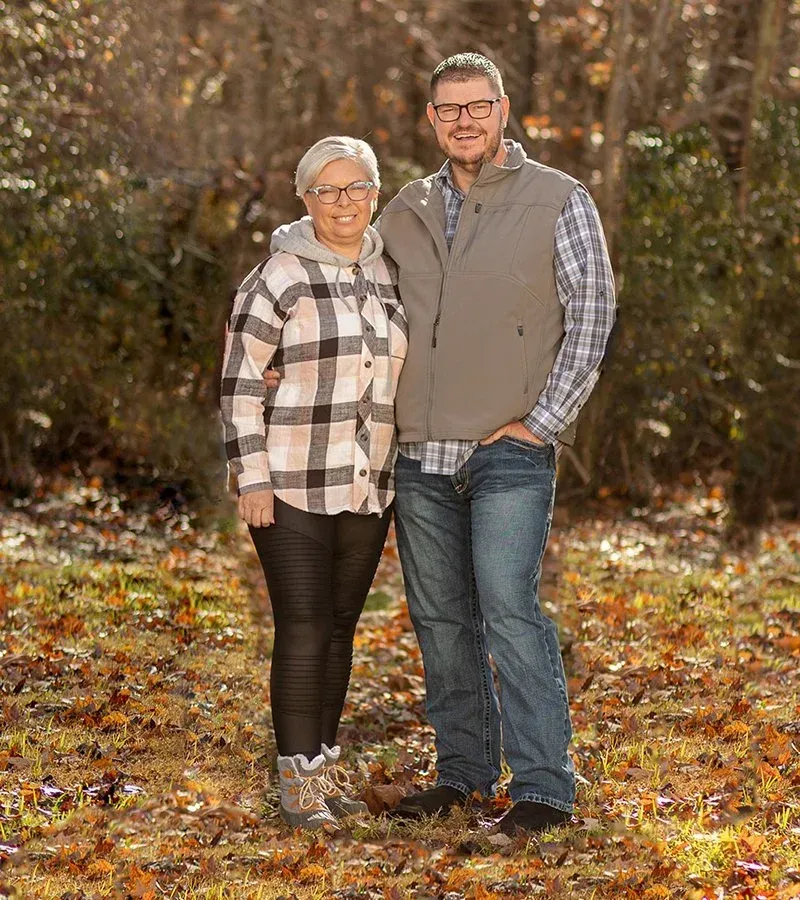 Couple standing outdoors. Woman wears plaid shirt, black leggings. Man wears vest, jeans. Fall foliage in background.