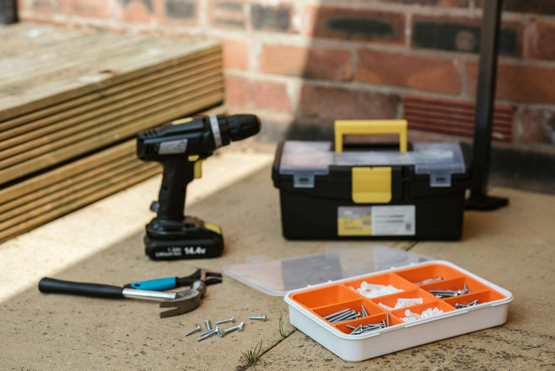 Tools on a concrete surface: drill, toolbox, hammer, screws, and an orange and white organizer, near a brick wall.