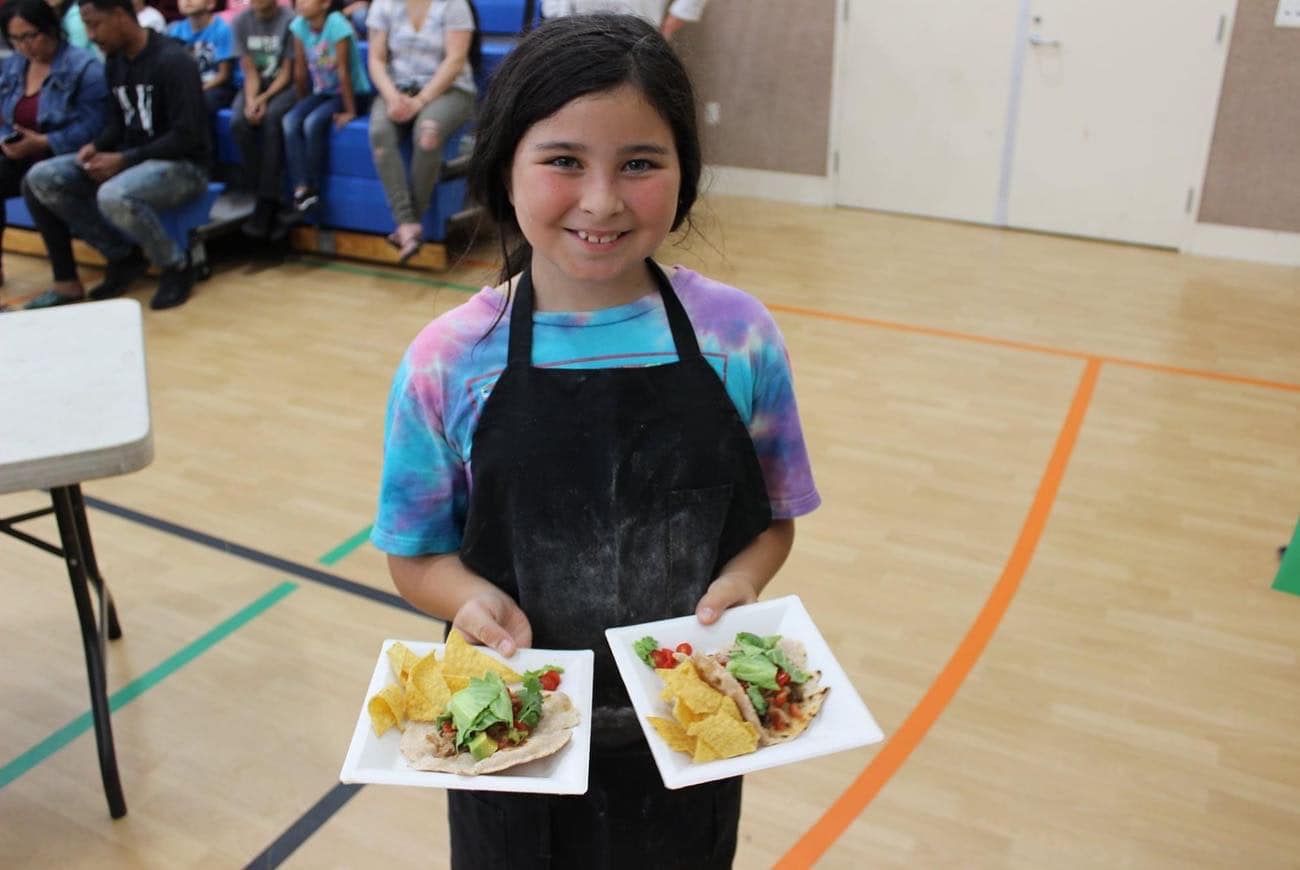 A young girl in an apron is holding two plates of food.