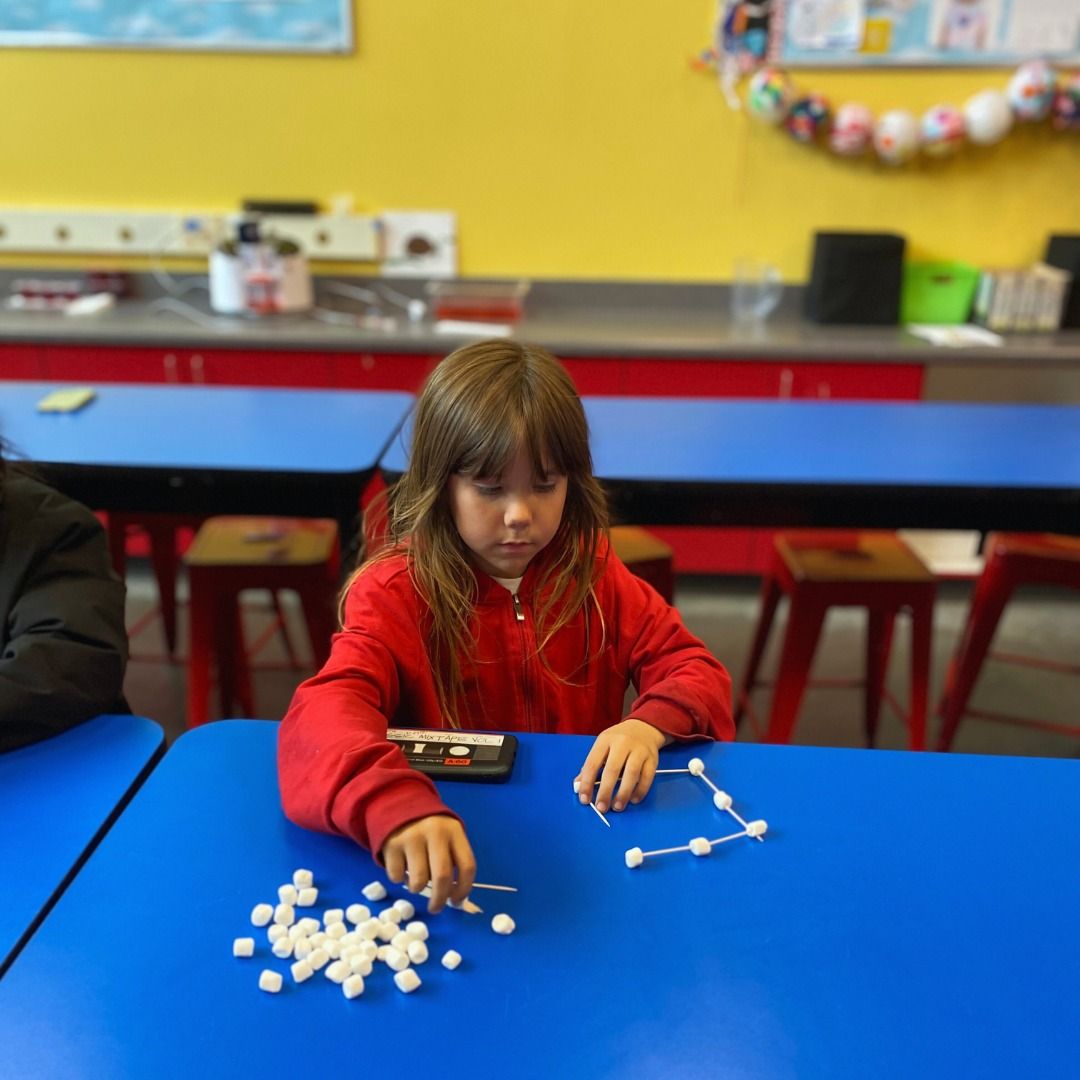 A little girl in a red jacket sits at a blue table