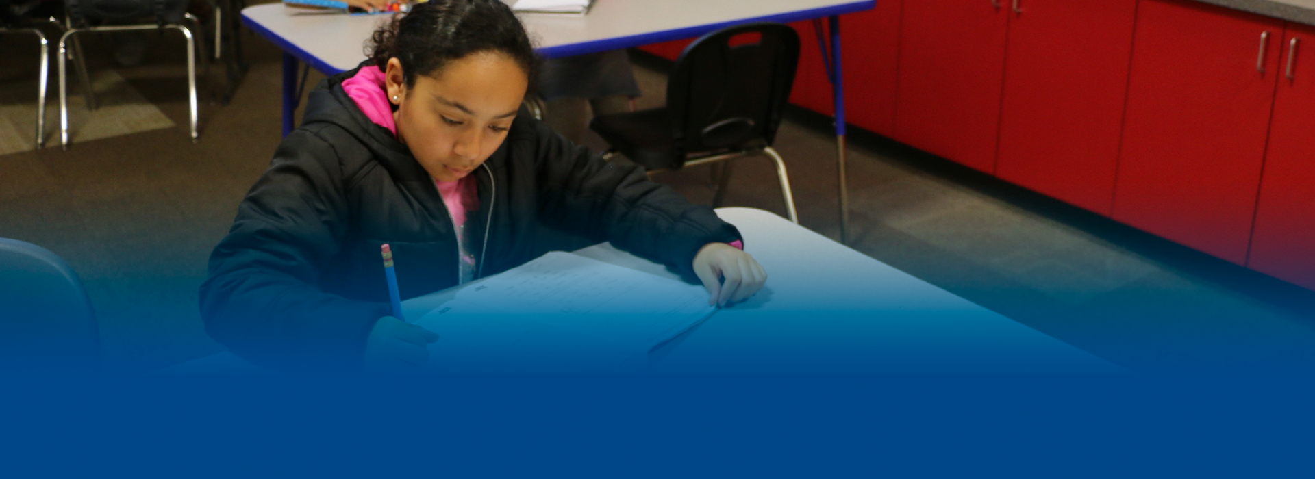 A young girl is sitting at a table in a classroom writing on a piece of paper.