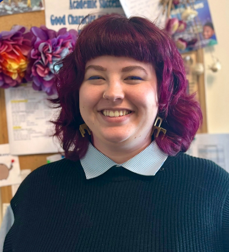 A woman wearing a striped shirt and earrings is smiling in front of a yellow and gray wall.