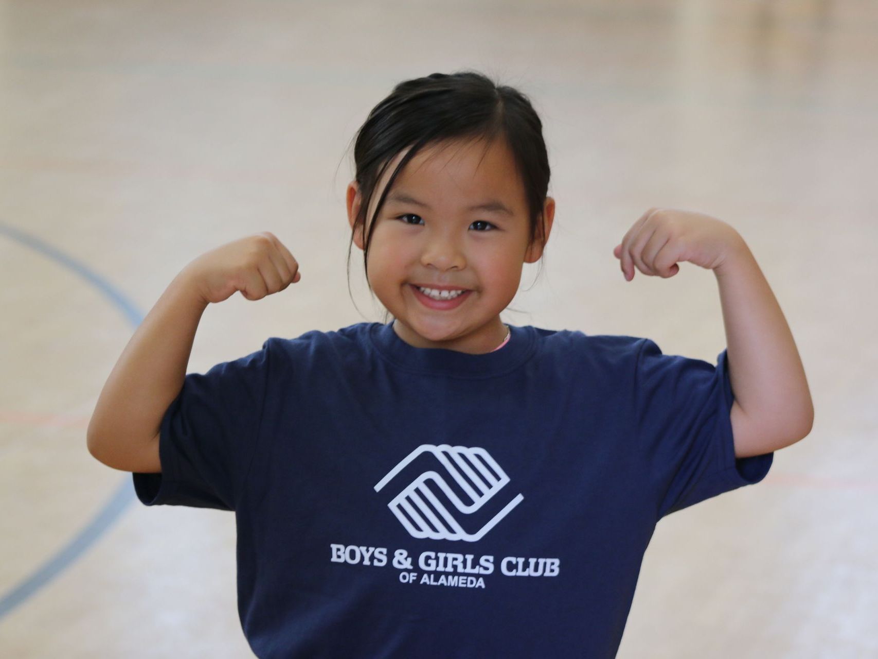 A young girl wearing a blue shirt that says boys and girls club of alaska flexes her muscles