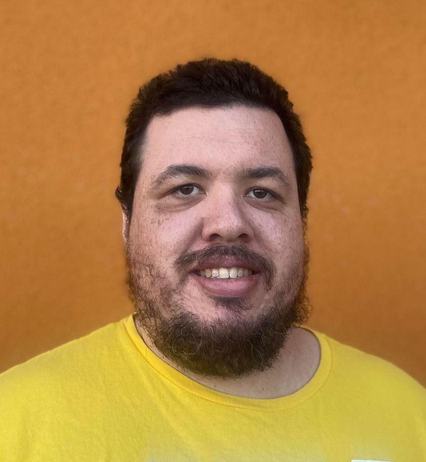 A man with curly hair and a beard is smiling in front of a yellow arrow.
