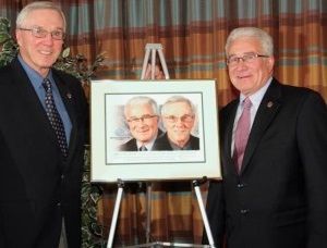 Two men in suits are standing next to a framed picture of two men