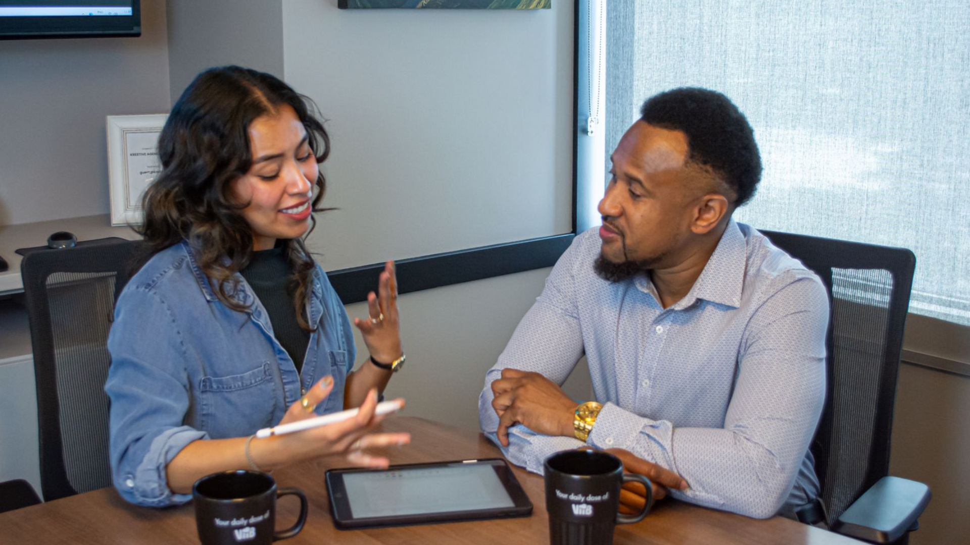 Two coworkers talking at a table with a tablet and coffee mugs in a conference room