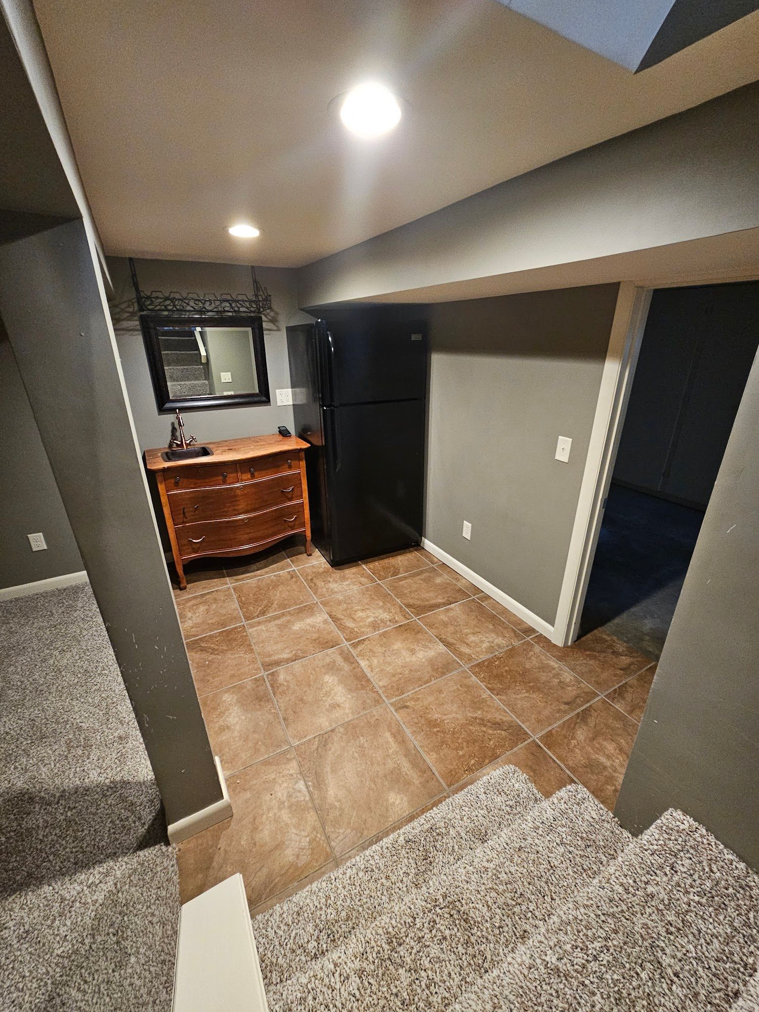 A basement with a refrigerator , dresser , mirror and stairs.