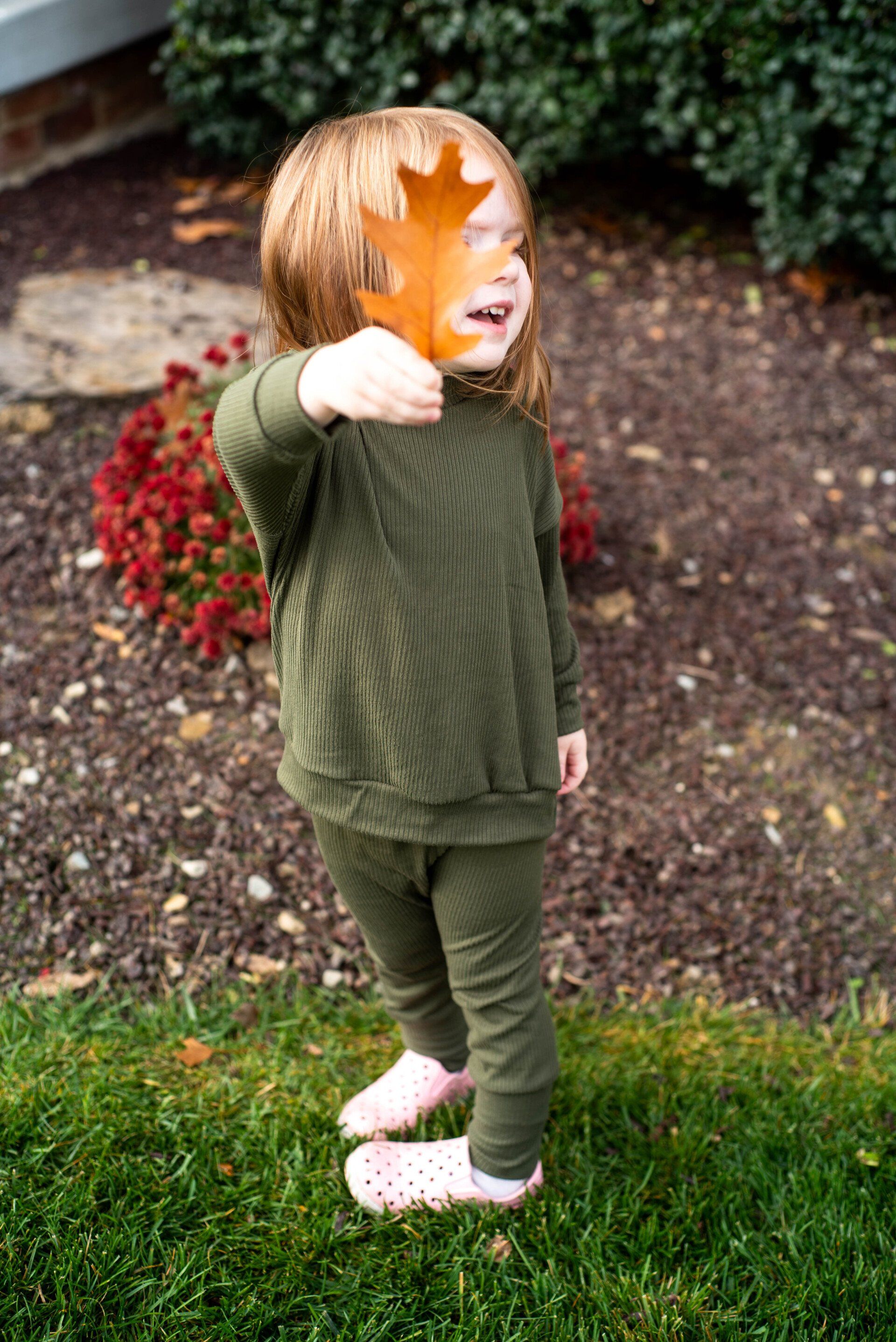 Little girl in a olive green shirt and pants holding up a orange oak leaf in front of her face.