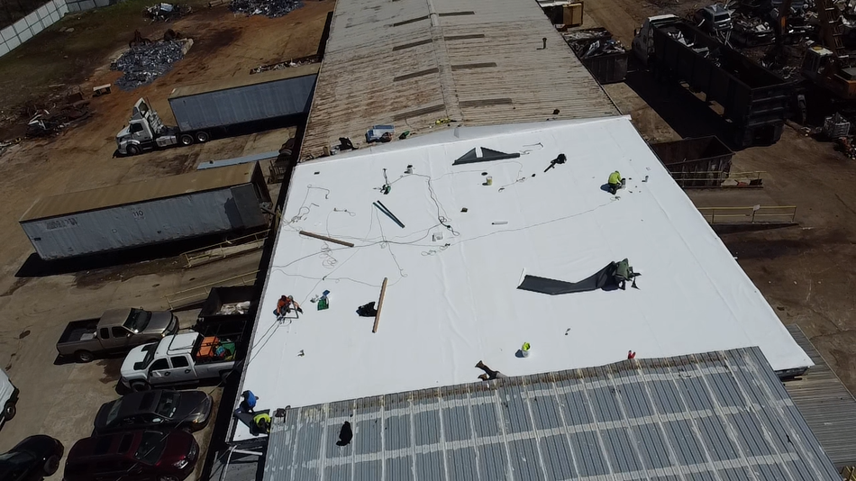 Aerial view of construction workers installing a white membrane roof on a large building at an industrial site.