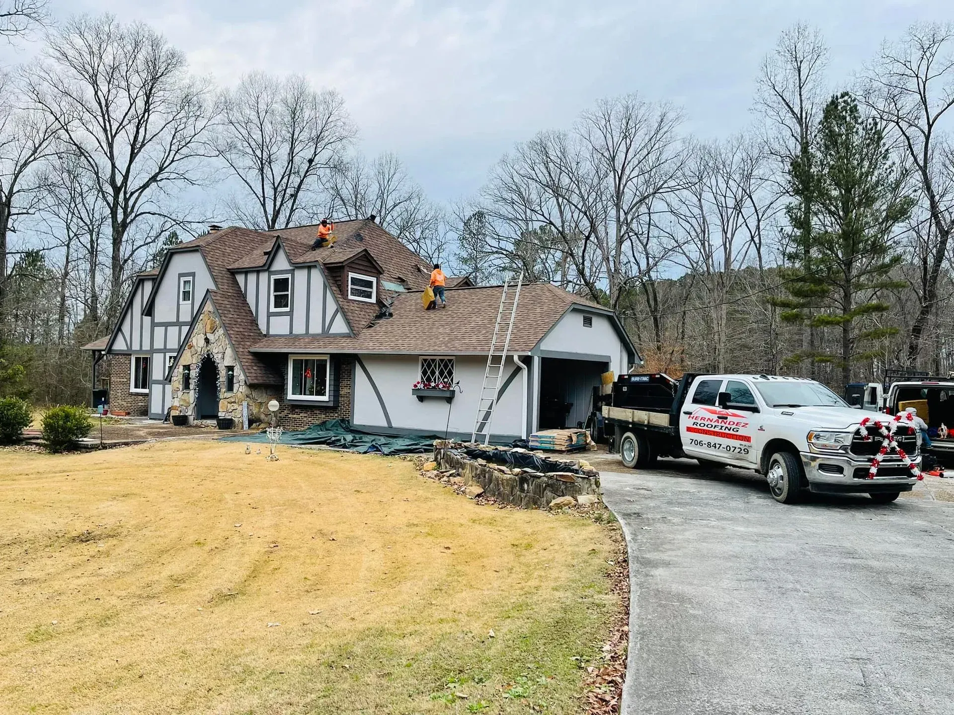 Workers repair a shingled roof on a two-story Tudor-style house with a white truck parked in the gravel driveway.