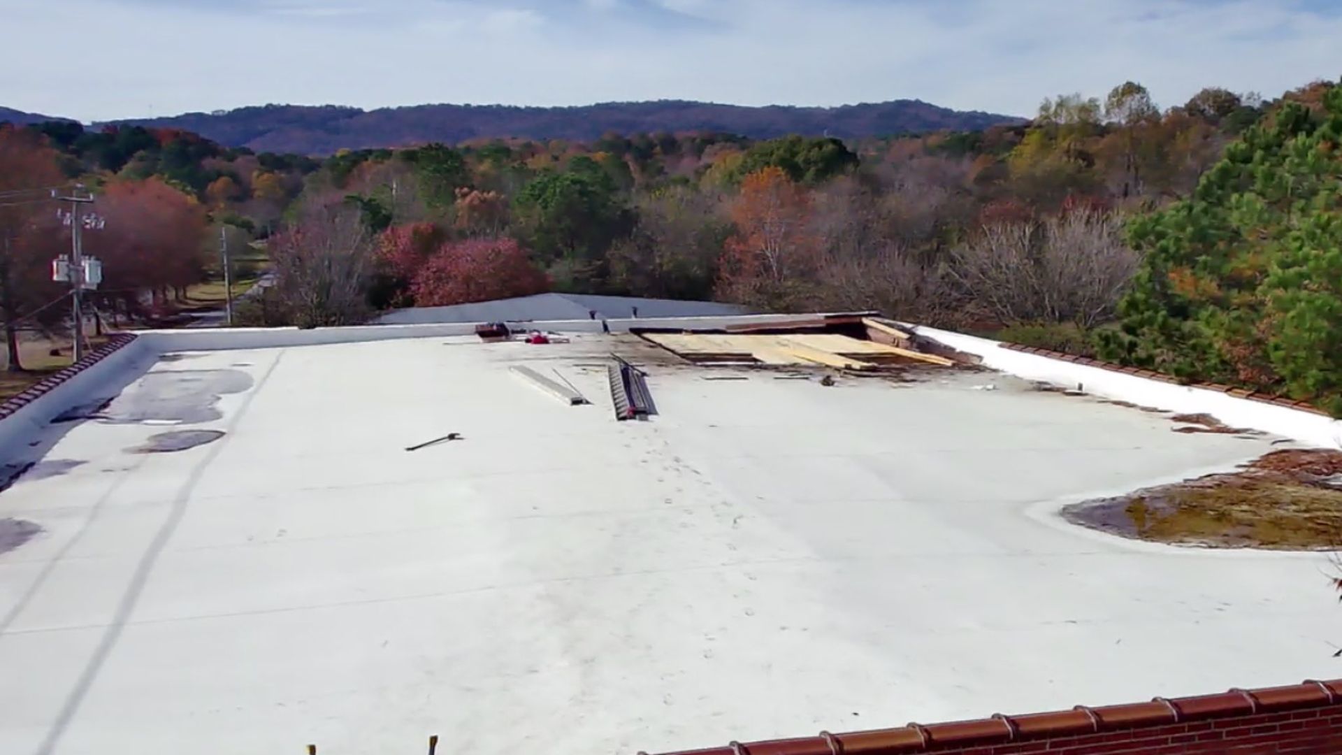 An elevated view of a large white flat roof under repair, with construction materials and debris, surrounded by trees.