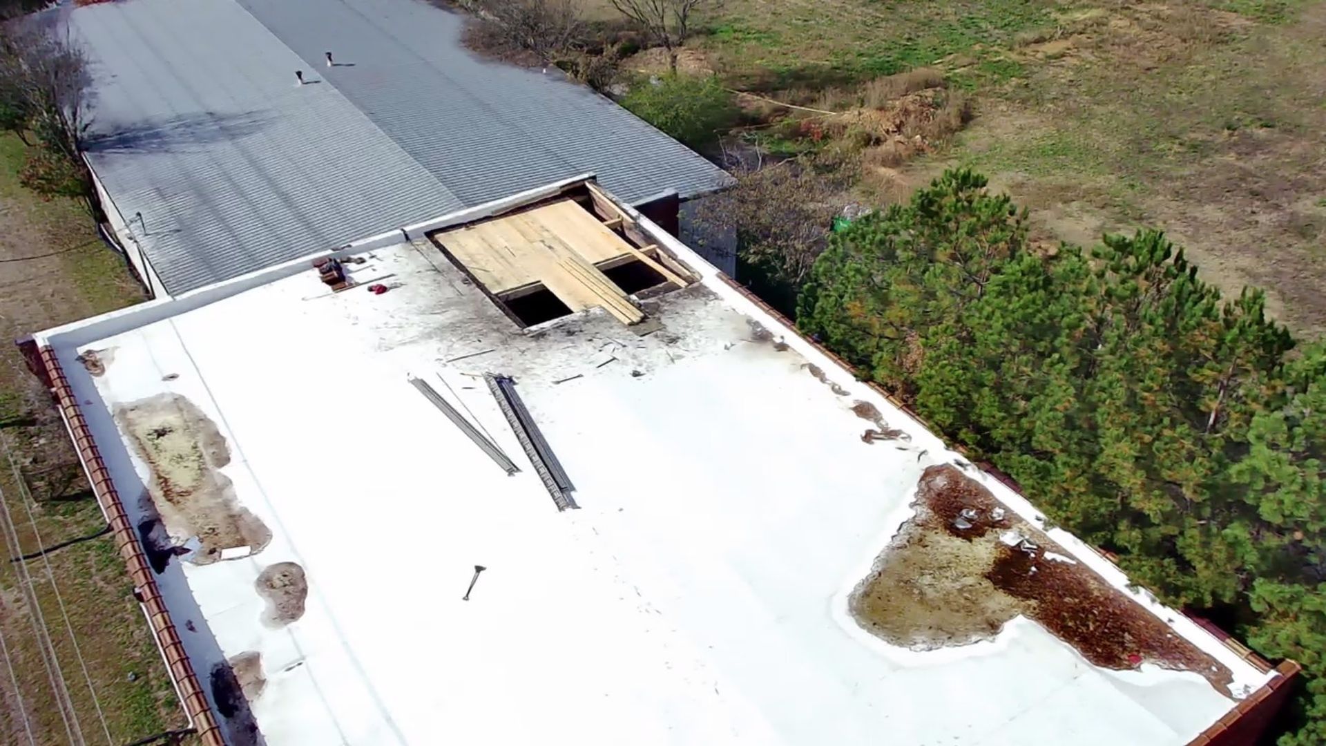 Aerial view of a flat commercial roof with damaged, patched sections and debris near a line of trees.